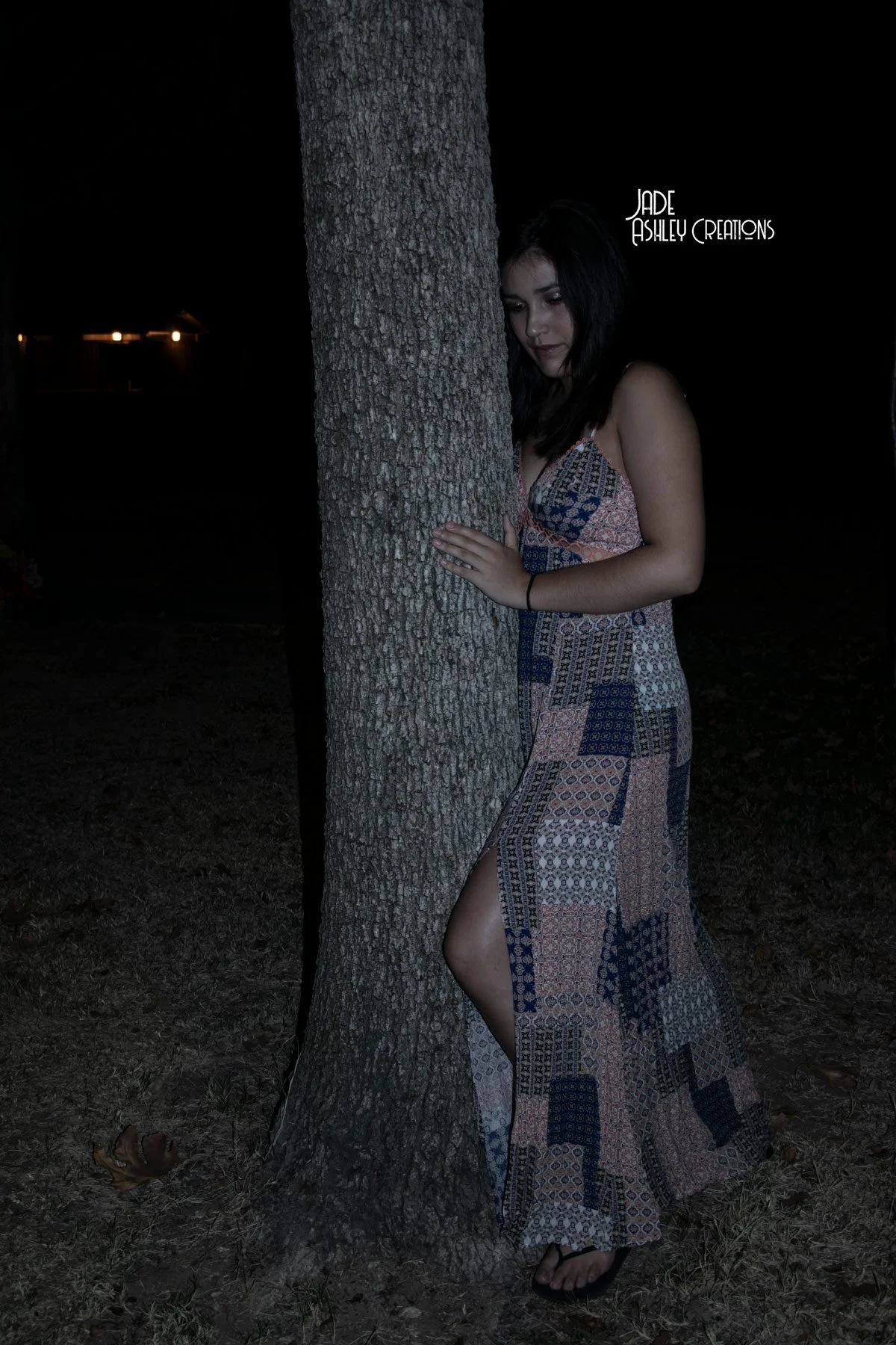 A woman in a patterned dress with a slit stands behind a tree at night, with her hands on the tree and her head slightly bowed, dark background, some distant lights.