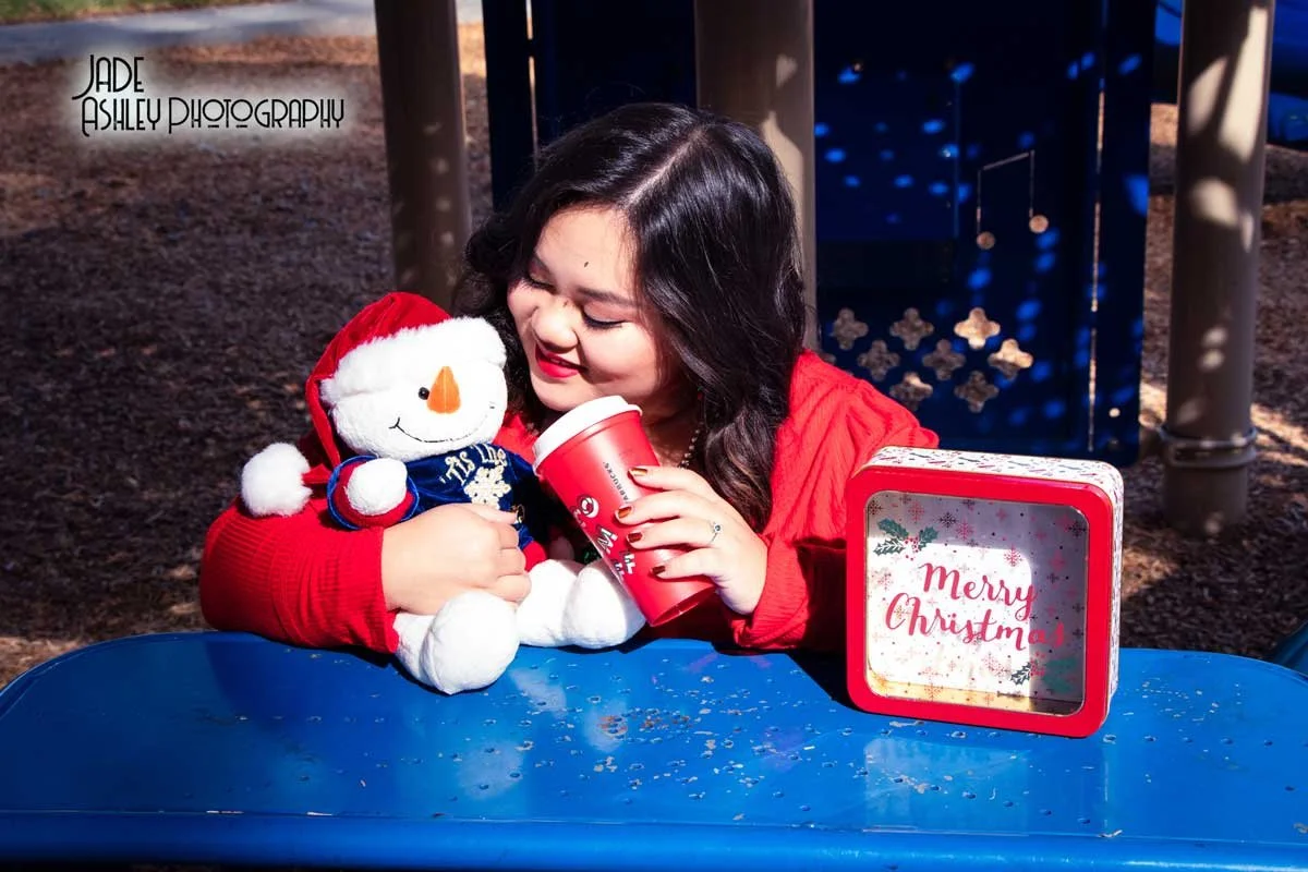 A young girl in a red jacket is smiling and hugging a plush snowman toy sitting on a blue picnic table. She holds a red cup in her hand and a 'Merry Christmas' decoration is on the table. Background includes playground equipment.