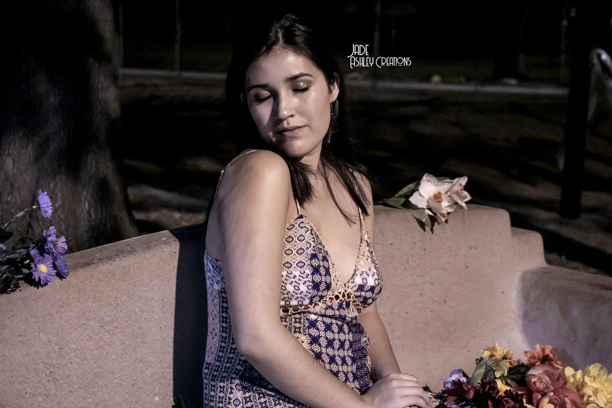 A young woman with dark hair and light skin sitting on a bench at night, wearing a floral dress, with purple and white flowers on the bench and in the background.