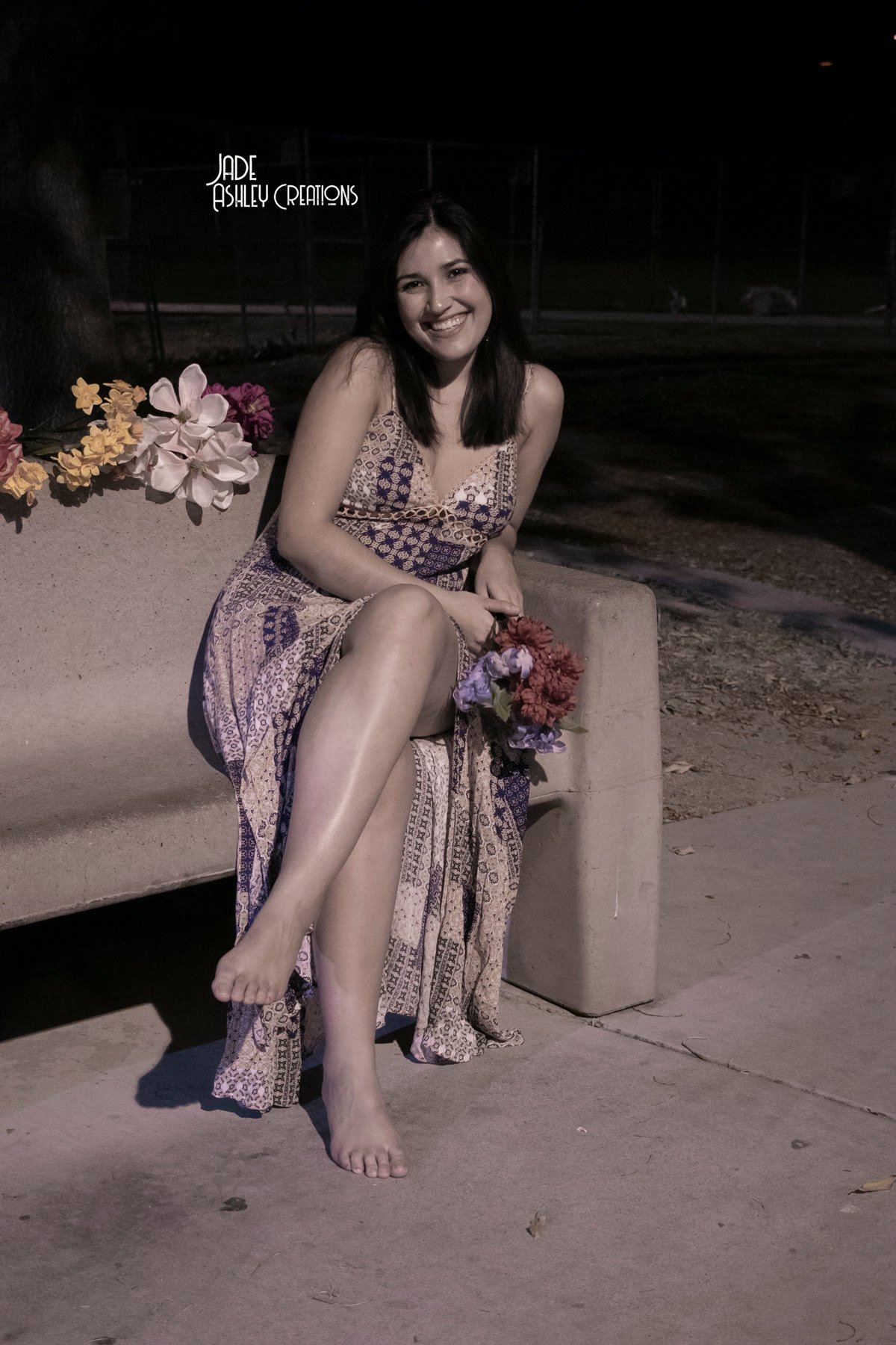 A young woman sitting on a concrete bench outdoors at night, smiling, holding a bouquet of flowers, wearing a patterned dress, with a dark background and some flowers visible on her left.