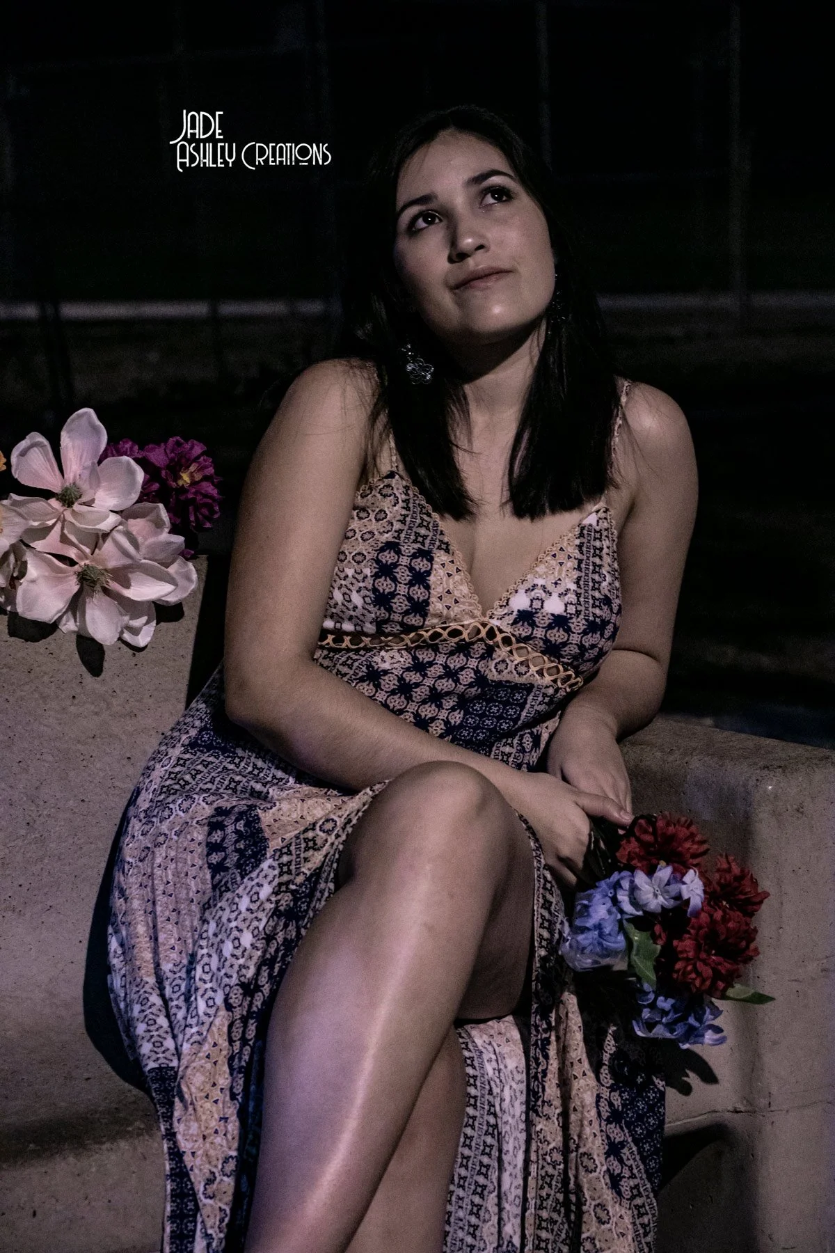 A woman with dark hair sits outdoors at night, holding a bouquet of flowers, with pink and purple flowers beside her, wearing a patterned dress.