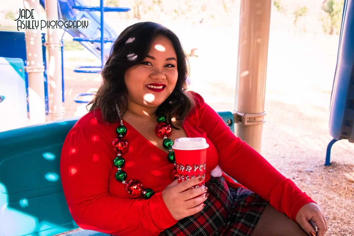 A woman in a red top and holiday necklace sitting on a blue bench holding a red cup with a holiday message, smiling at the camera. She is outdoors near a playground.