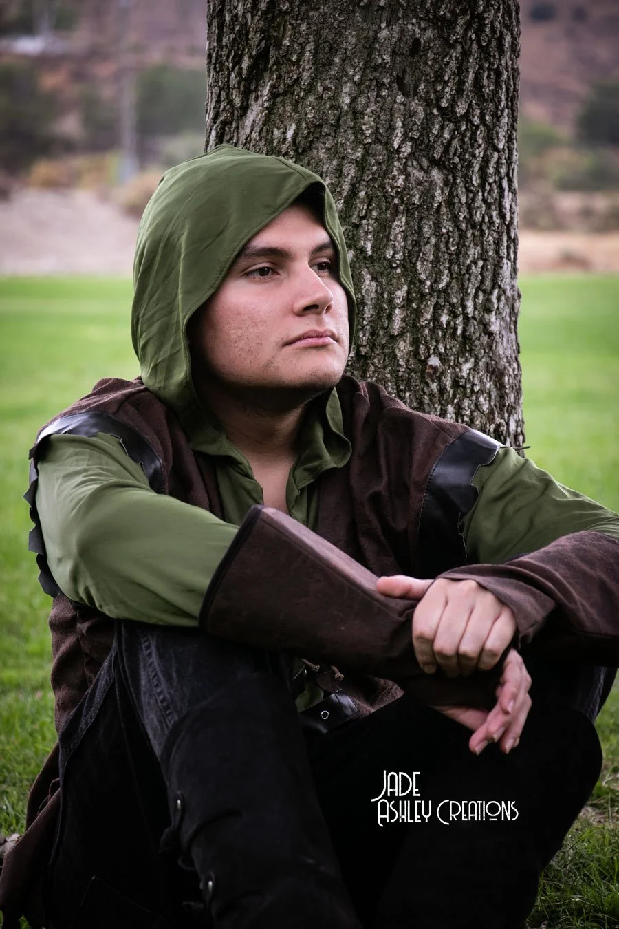 A young man wearing a green hooded jacket sitting against a tree in a park, looking contemplative.
