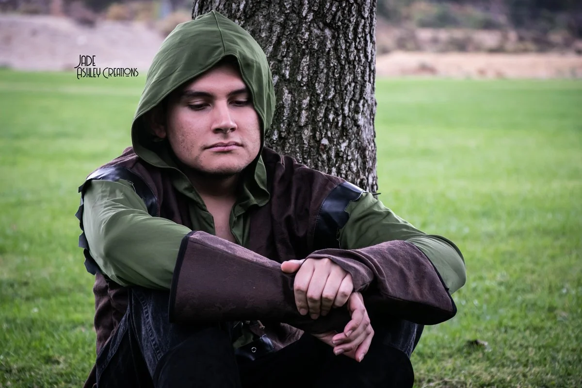 A young man sitting on the grass near a tree with his eyes closed, wearing a green hoodie with the hood up and brown sleeves, with a contemplative or peaceful expression.