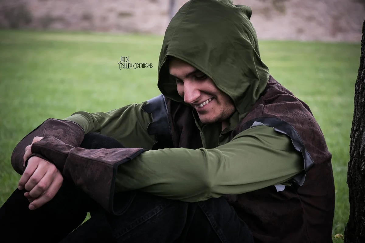 A young man sitting outdoors on the grass, smiling and looking down, wearing a green hoodie and brown jacket.