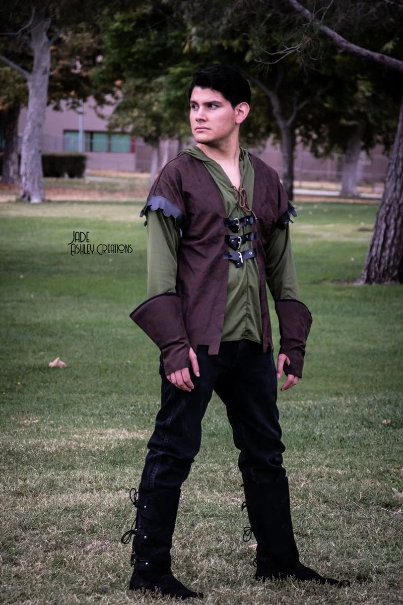 A young man with dark hair standing outdoors in a park-like setting, dressed in medieval or fantasy-inspired costume with green and brown clothing and tall black boots.