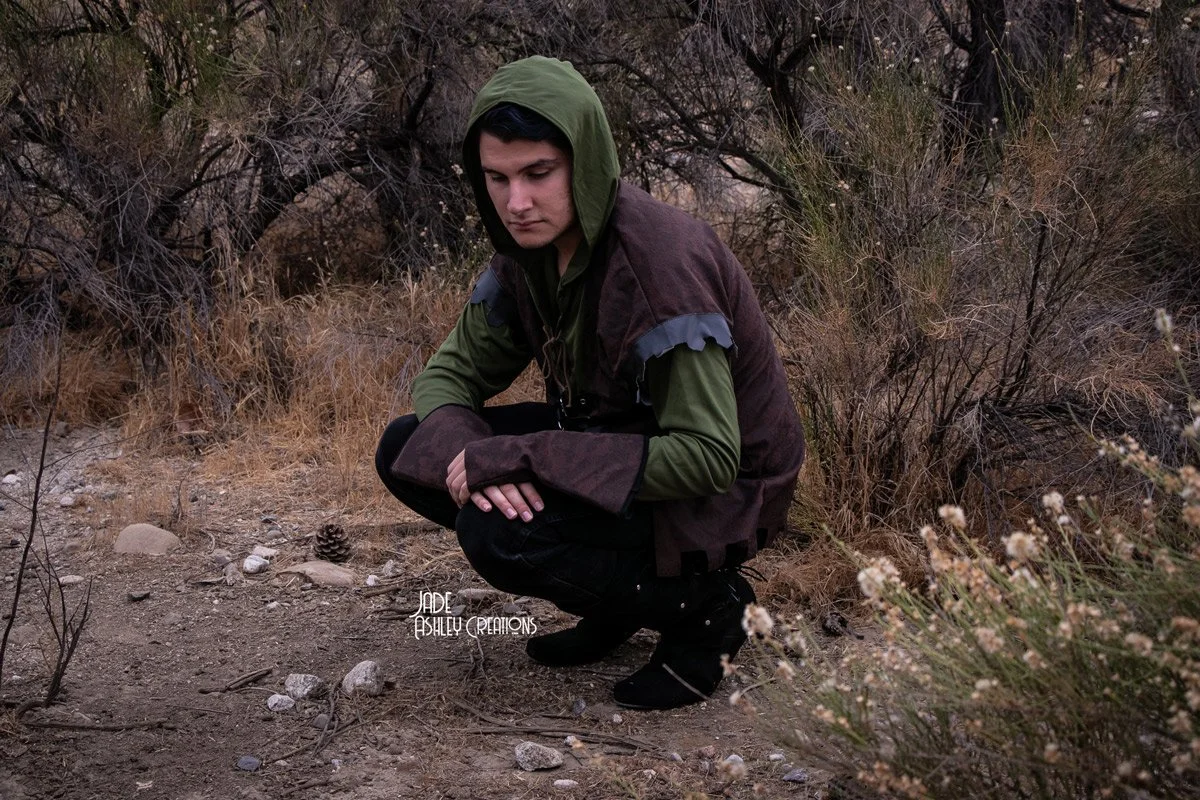 A young man wearing a green hoodie and brown jacket crouches on a dry, rocky trail surrounded by sparse bushes and trees.