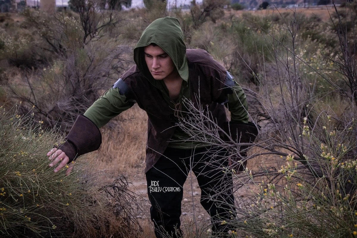 A young man wearing a hooded jacket and dark pants, crouching in a dry, brushy outdoor landscape with sparse bushes and trees.