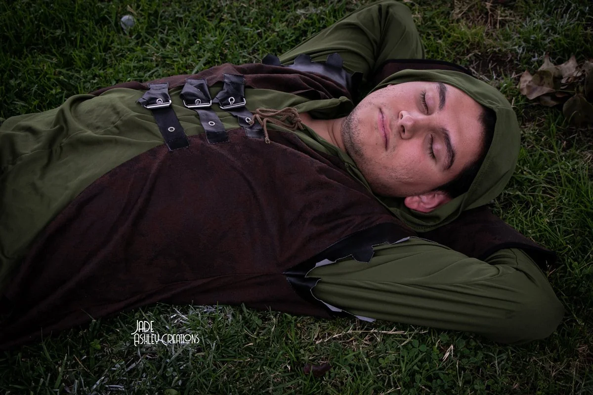 A man dressed in a green hooded cloak and brown tunic, lying on the grass with his eyes closed, as if sleeping outdoors.