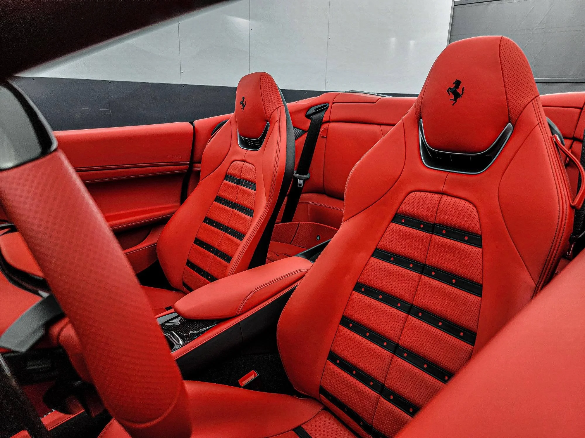 Red Ferrari car interior with leather seats, seat belts, and the iconic prancing horse logo embroidered on the headrests.