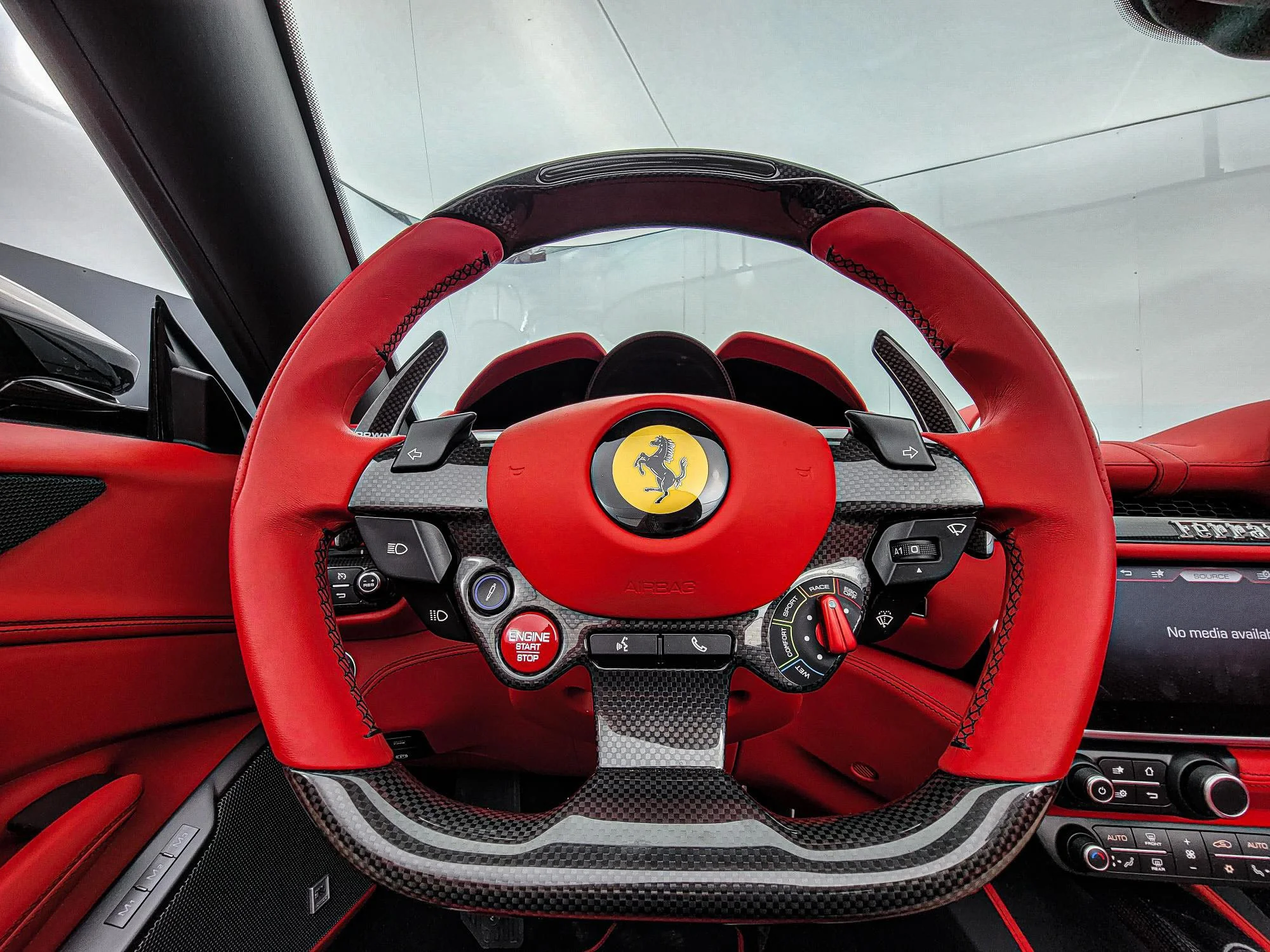 Close-up of a red and black Ferrari interior, focusing on the steering wheel with the Ferrari logo. The steering wheel is equipped with various controls, a red start/stop button, and a combination of carbon fiber and leather materials.