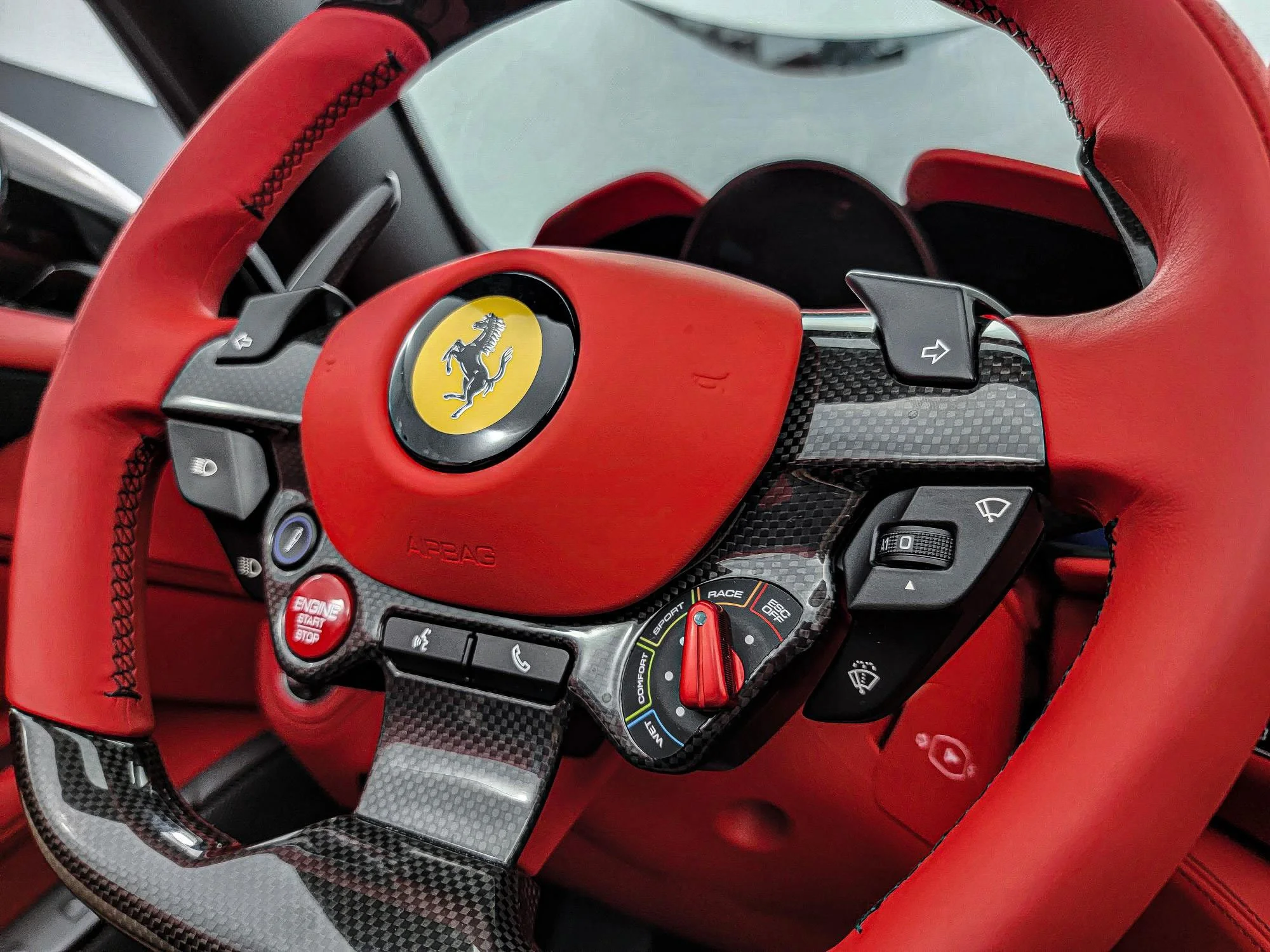 A close-up of a Ferrari racing car steering wheel with the Ferrari logo, various control buttons, and a carbon fiber finish.