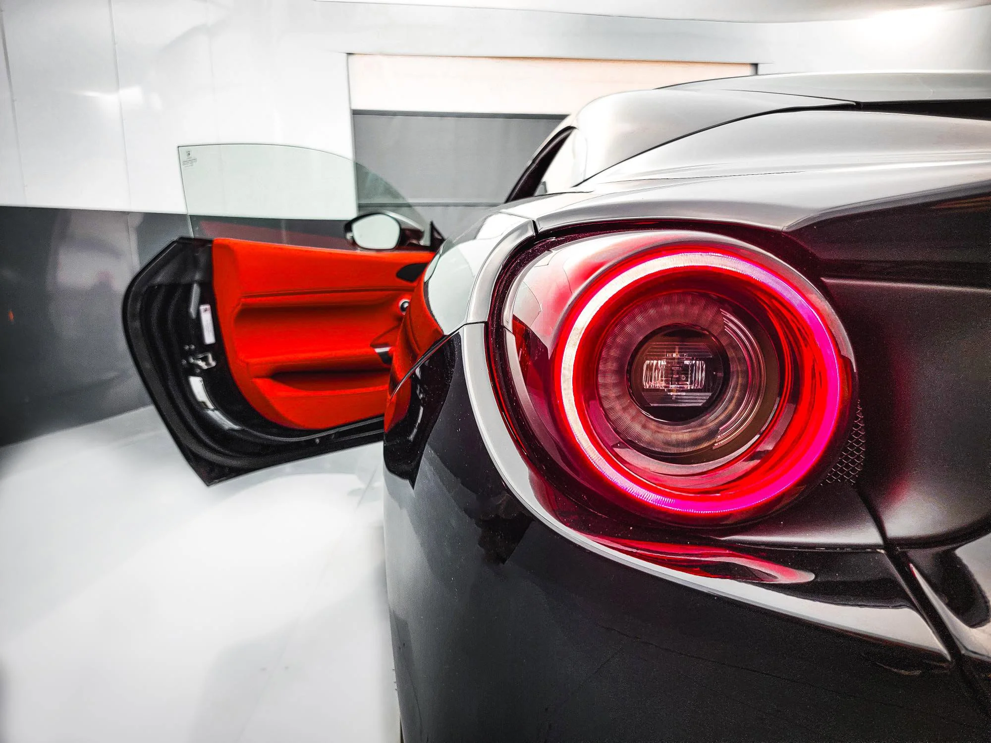 Close-up of a sleek black sports car with a glowing red tail light, set against a modern, minimalistic white and gray background, with the driver side door open revealing a red interior.