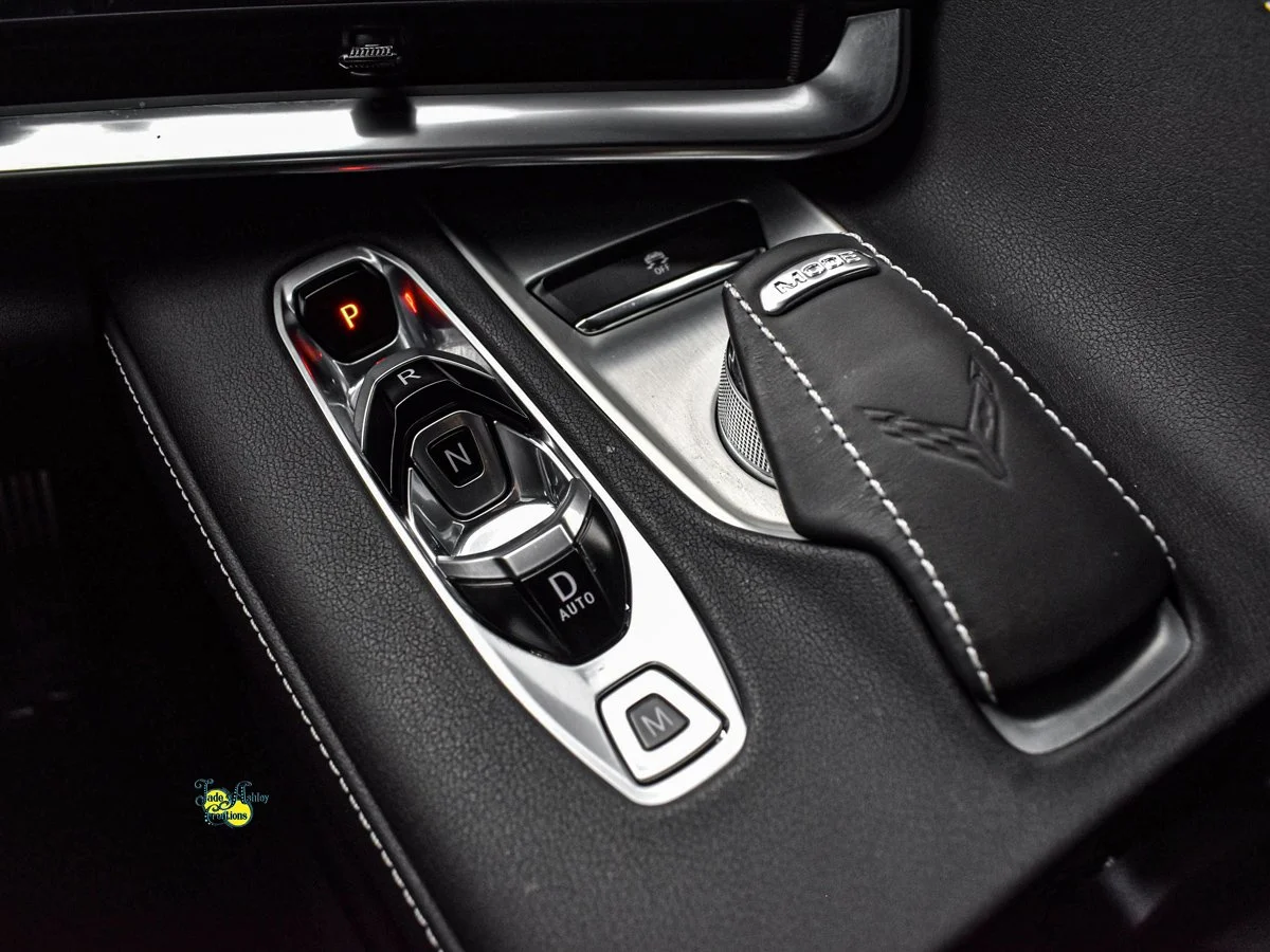 Close-up of a car gear shift with P, R, N, D auto, and M modes, and a leather shift cover with Corvette logo.