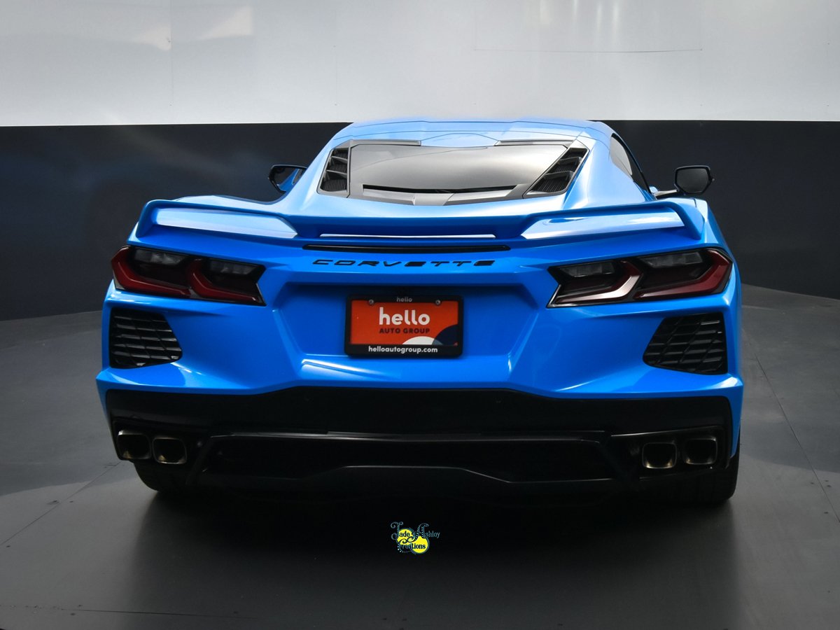 Blue Chevrolet Corvette sports car displayed indoors with a black and gray background, showing the rear view with an orange 'hello AUTO GROUP' dealer plate.