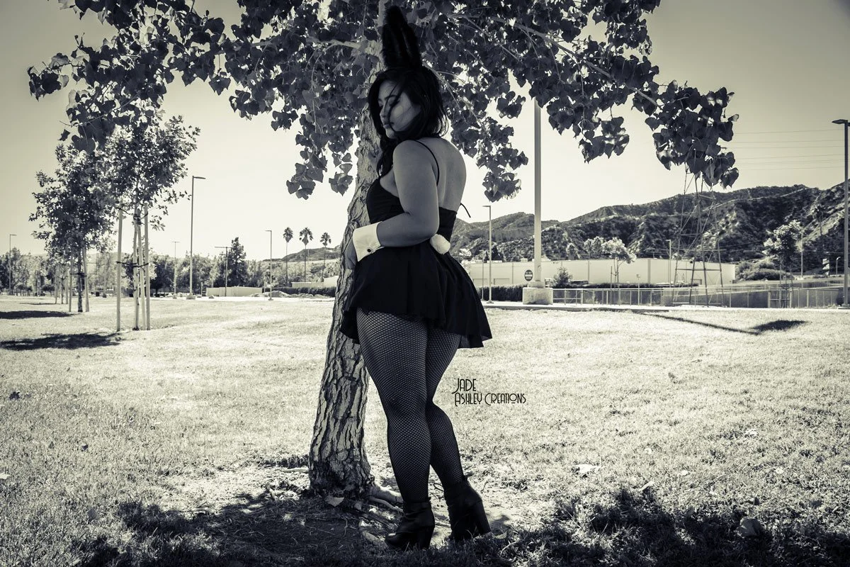 A woman dressed in a bunny costume with a black dress, fishnet stockings, high heels, and bunny ears, standing outdoors under a tree, with hills and a park in the background, in black and white.