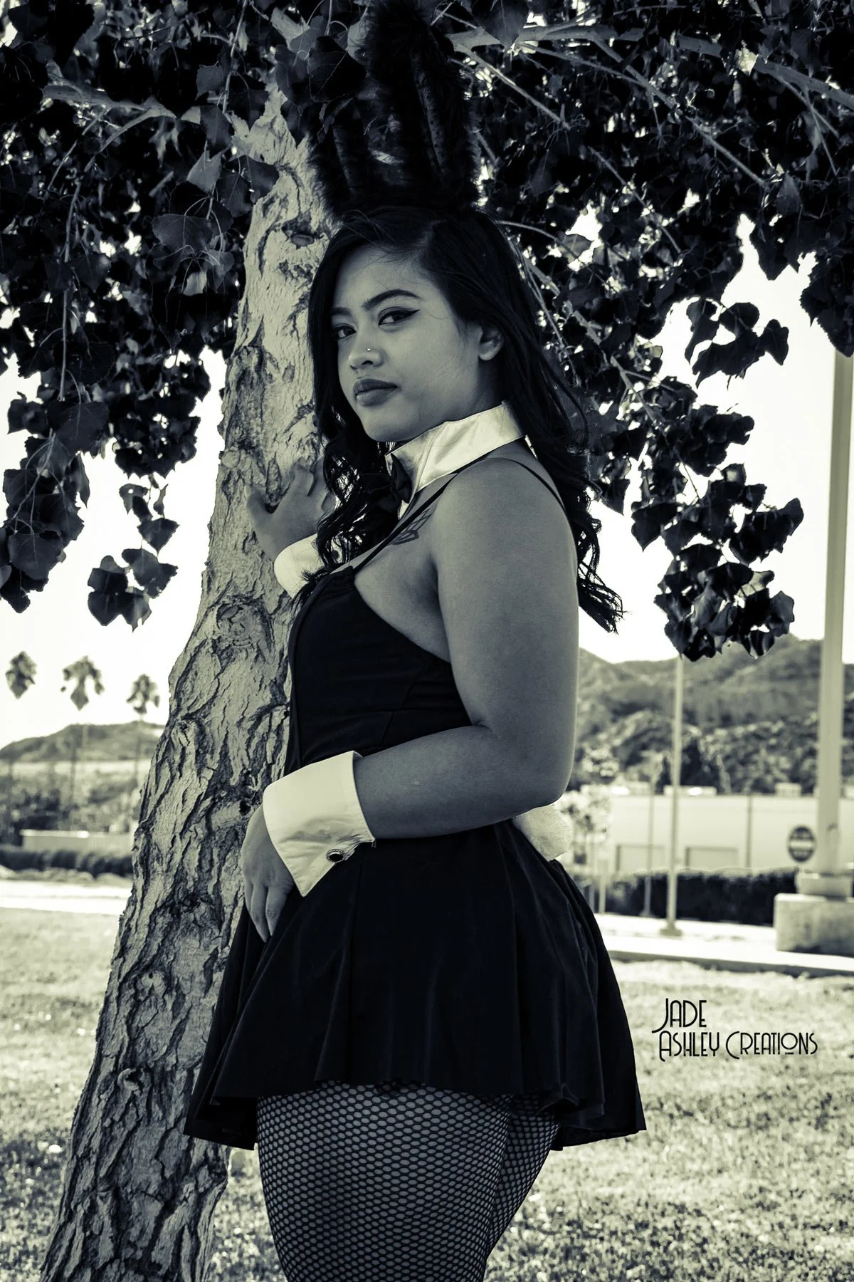 A woman dressed as a sexy waitress with a black dress, fishnet stockings, and a white collar with a bow tie, standing outdoors next to a tree with leaves and a background of mountains and a parking lot.