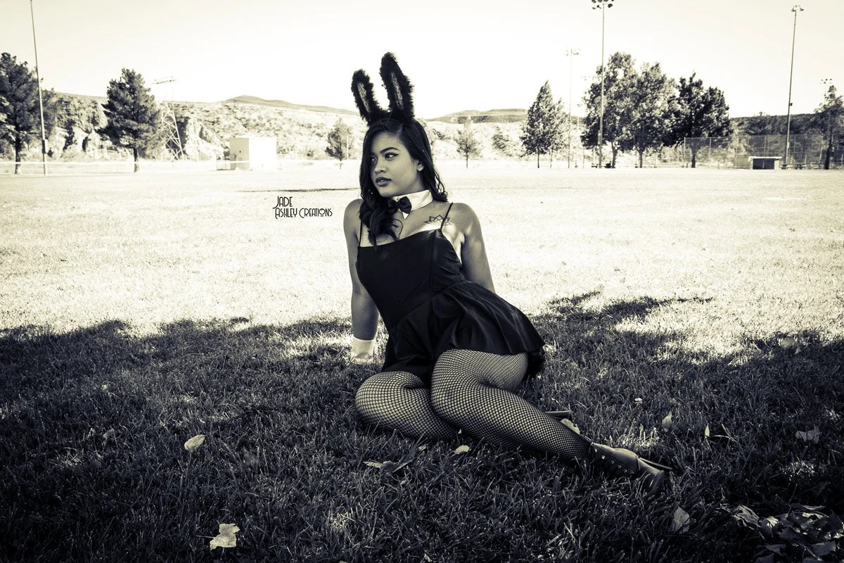 A woman dressed in a bunny girl costume with fishnet stockings, sitting on grass in an open field with trees and hills in the background.