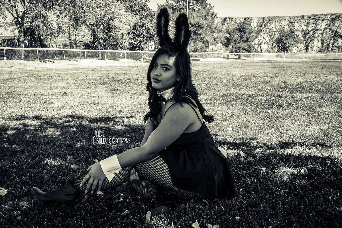 Young woman sitting on grass outdoors wearing bunny ears, fishnet stockings, a black dress, and boots in a black-and-white photo.