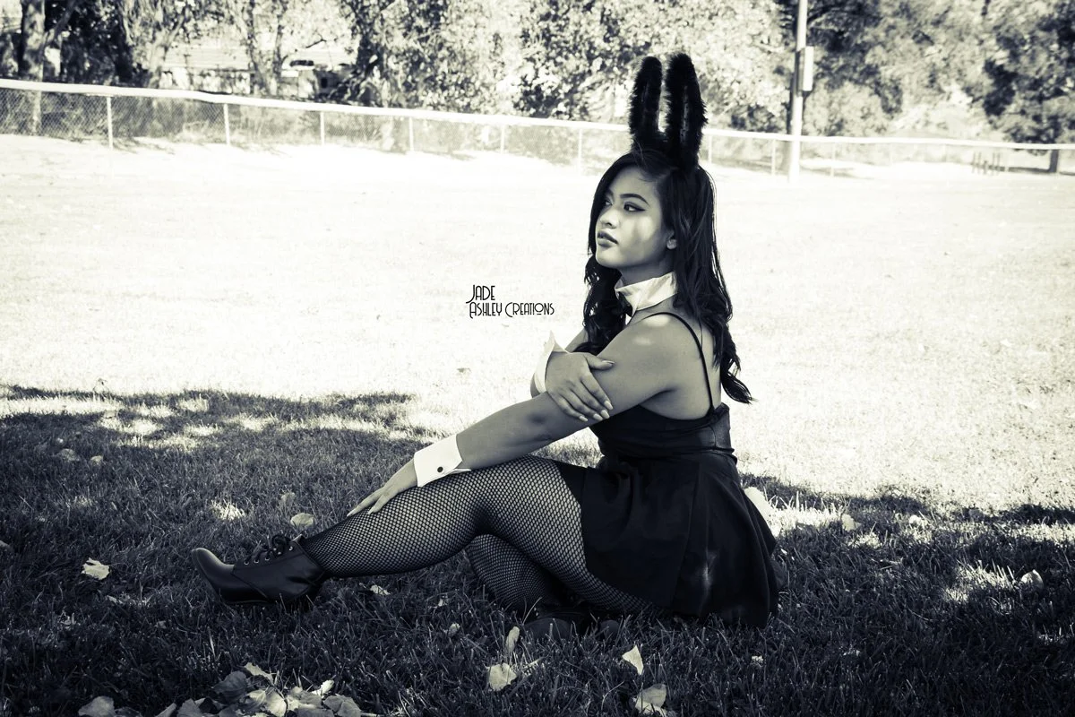 A woman dressed in a French maid costume with bunny ears, sitting on grass in a park, black and white photo.