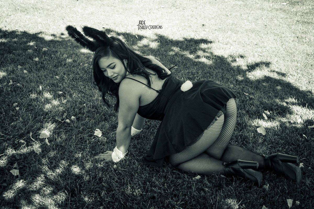 A woman dressed in a bunny costume with bunny ears, fishnet stockings, and high heels, posing on her hands and knees on grass in a park.