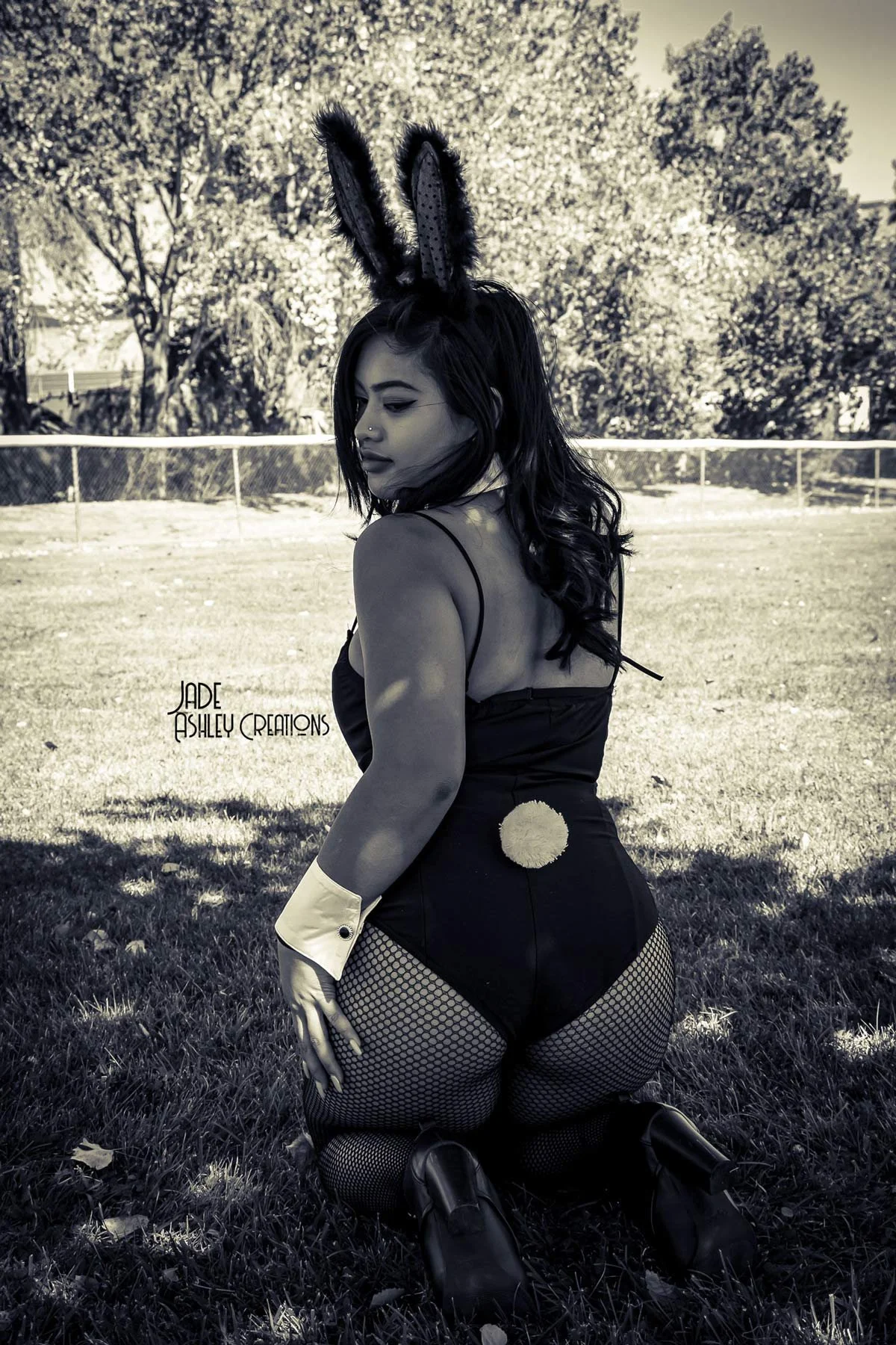 A woman dressed in a bunny costume with bunny ears, fishnet stockings, black shoes, and a tail, kneeling on grass in an outdoor park with trees and a fence in the background.