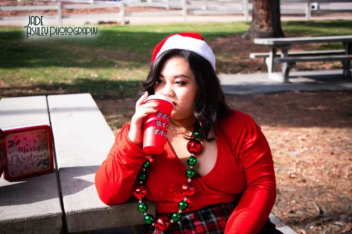 A woman dressed in red, wearing a Santa hat and holiday jewelry, sitting outdoors and drinking from a red cup. There is a small Christmas sign on the table next to her, and a park setting with grass, a tree, and picnic tables in the background.