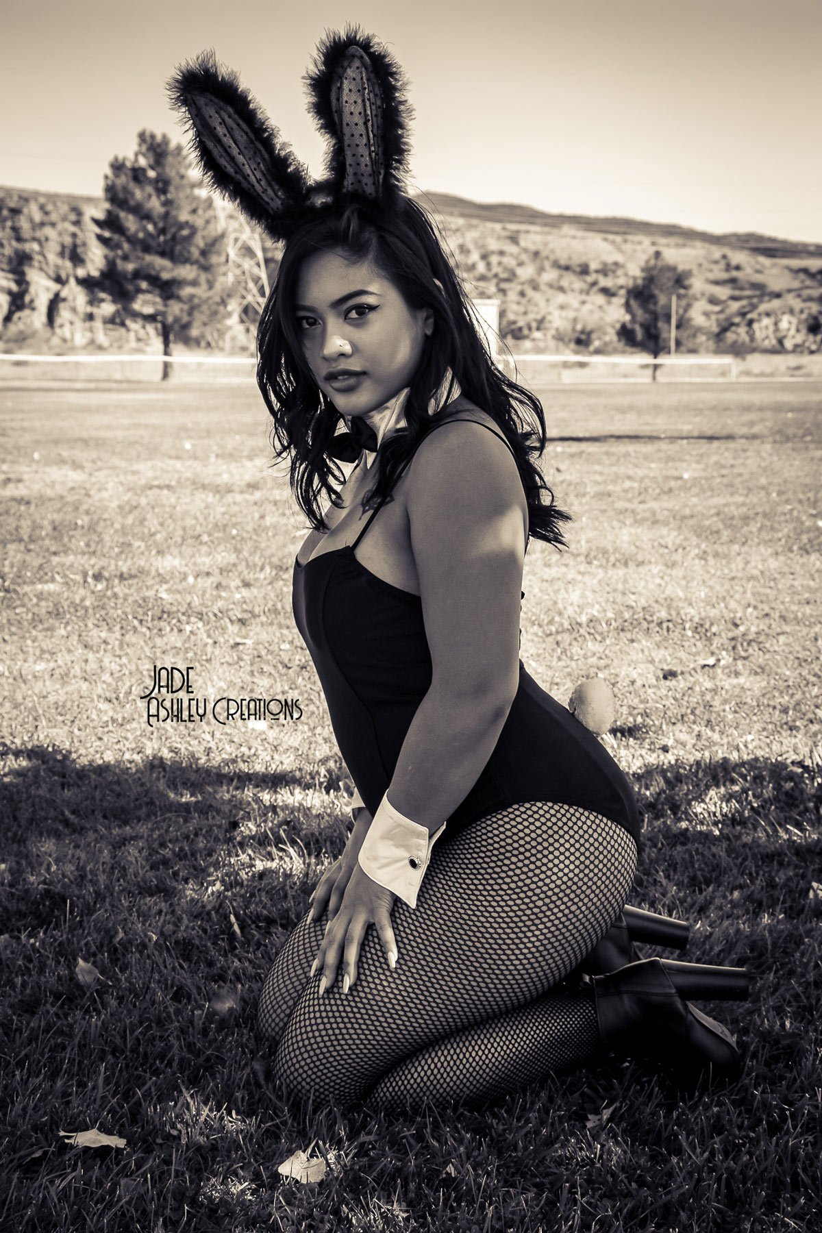 A woman dressed in a bunny costume with bunny ears, fishnet stockings, and high heels, kneeling on grass outdoors with mountains and trees in the background.