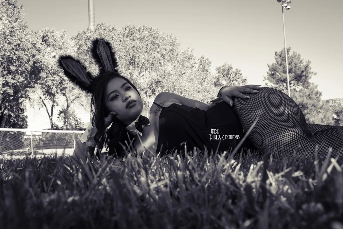 A woman lying on the grass outdoors, wearing bunny ears, a black outfit, fishnet stockings, and a bow tie, with trees and a fence in the background.