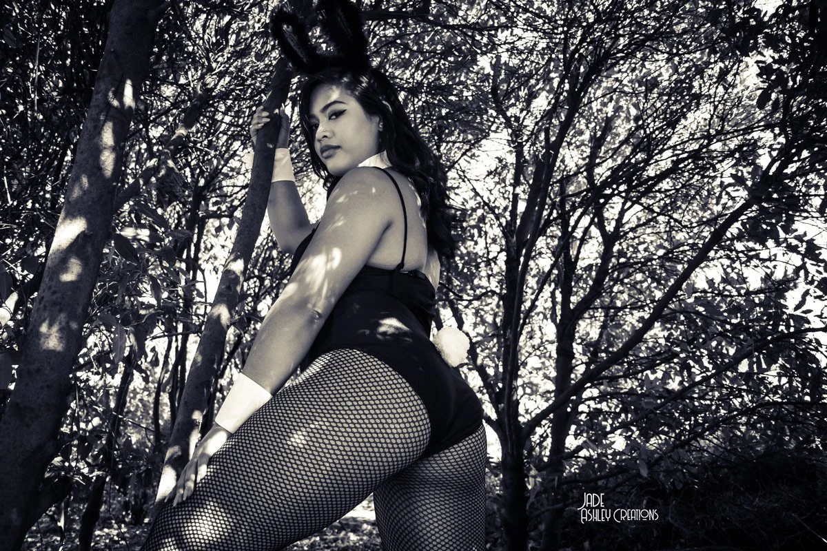 A woman dressed in a bunny costume with fishnet stockings, standing outdoors among trees, looking over her shoulder at the camera.