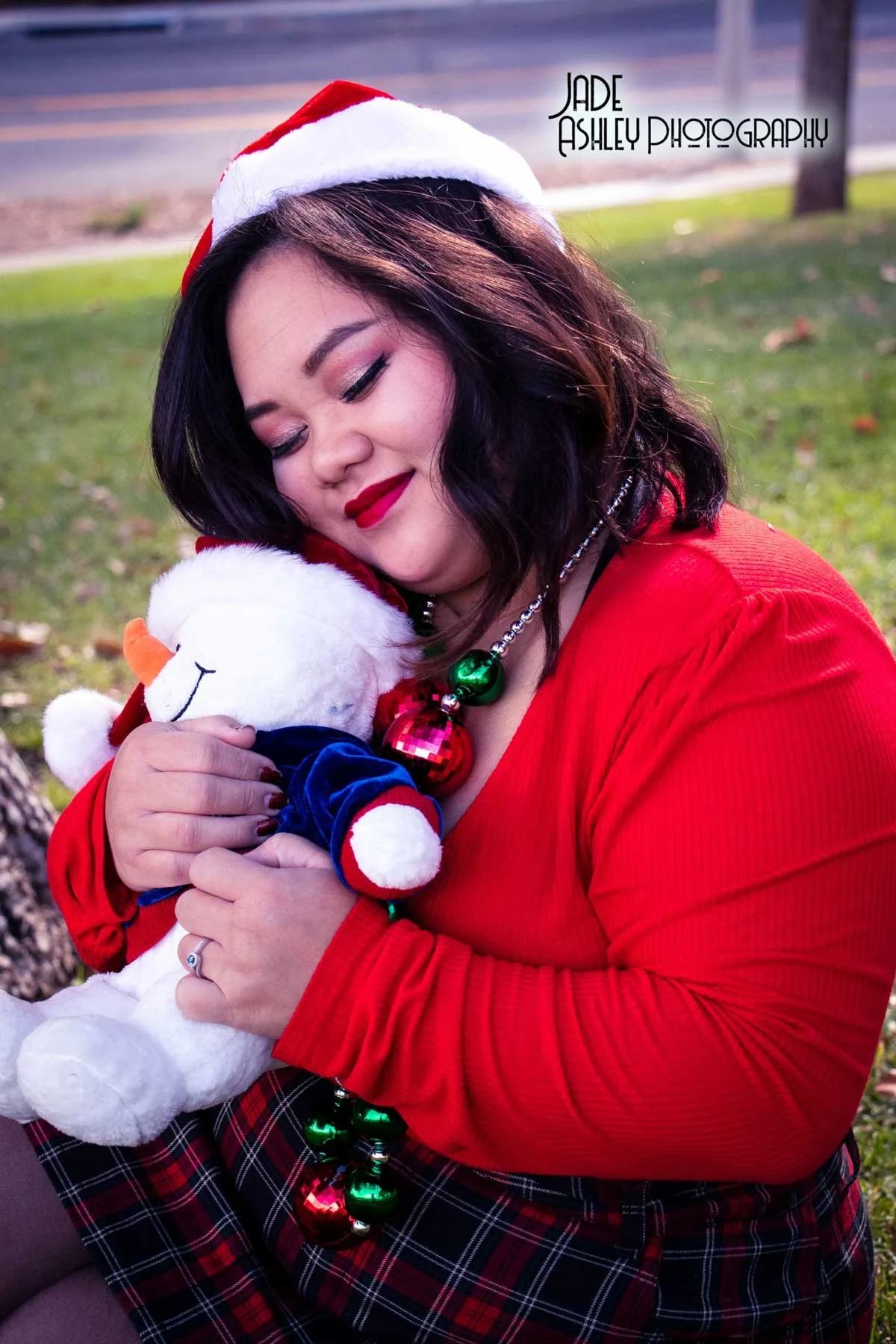 A woman with dark hair and red lipstick wearing a red Santa hat and red top, hugging a white plush snowman toy dressed in a Santa outfit. She is outdoors with a grassy background and a blurred road in the distance.