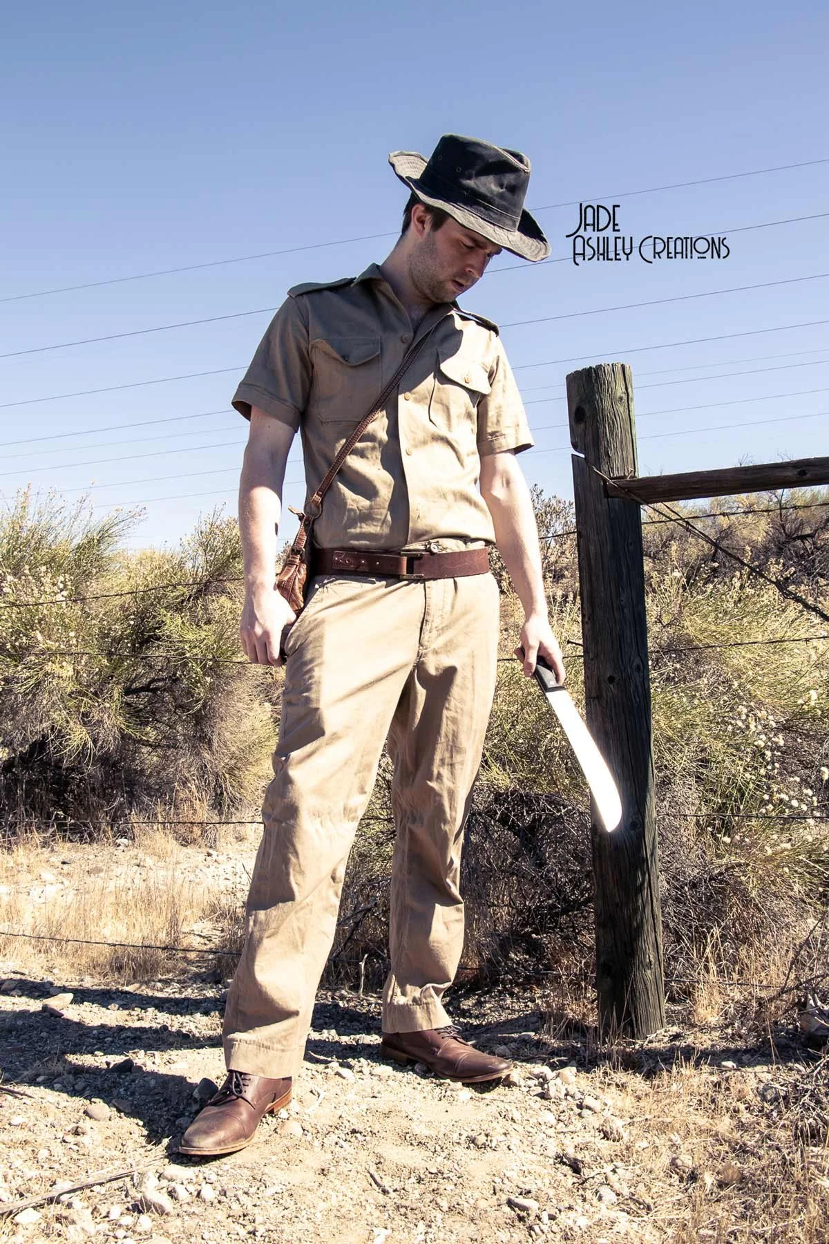 A man dressed as a park ranger holding a machete, standing outdoors on dry terrain with shrubbery and a wooden fence post visible in the background.