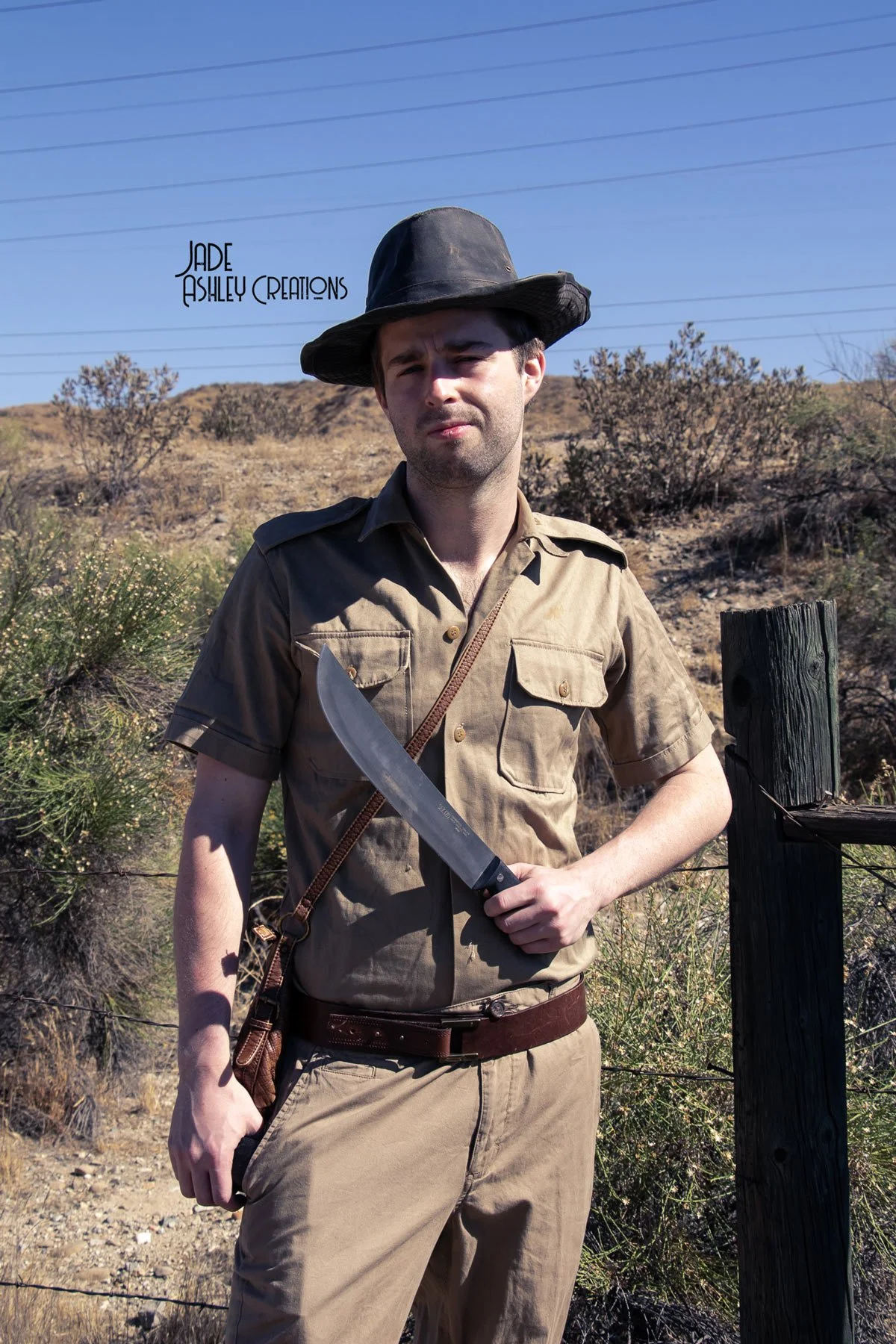 A man dressed as a park ranger holding a large knife in a desert landscape with dry bushes and a wooden post.