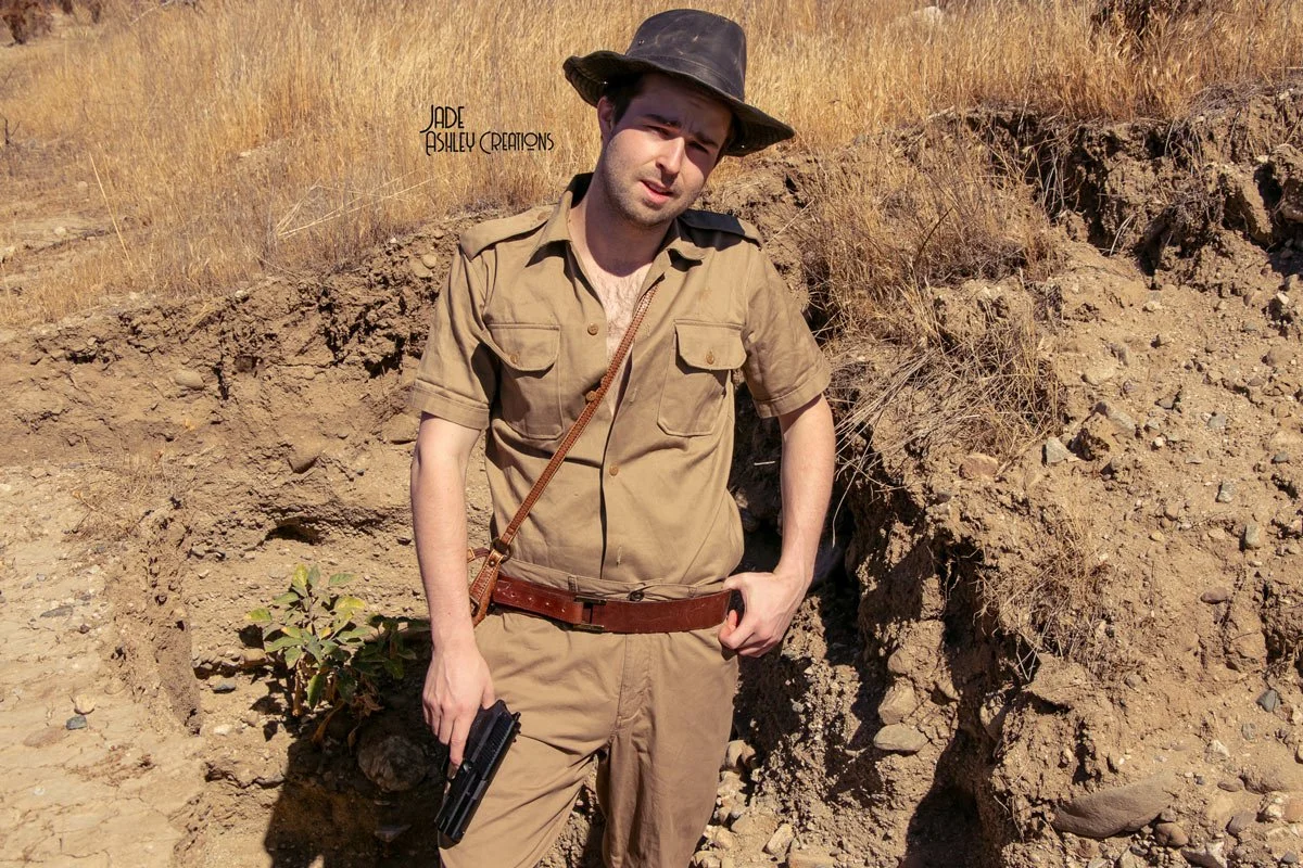 A man dressed as an explorer or adventurer, wearing a khaki outfit, wide-brimmed hat, and holding a handgun, standing next to a hole or excavation site in a barren, grassy landscape.