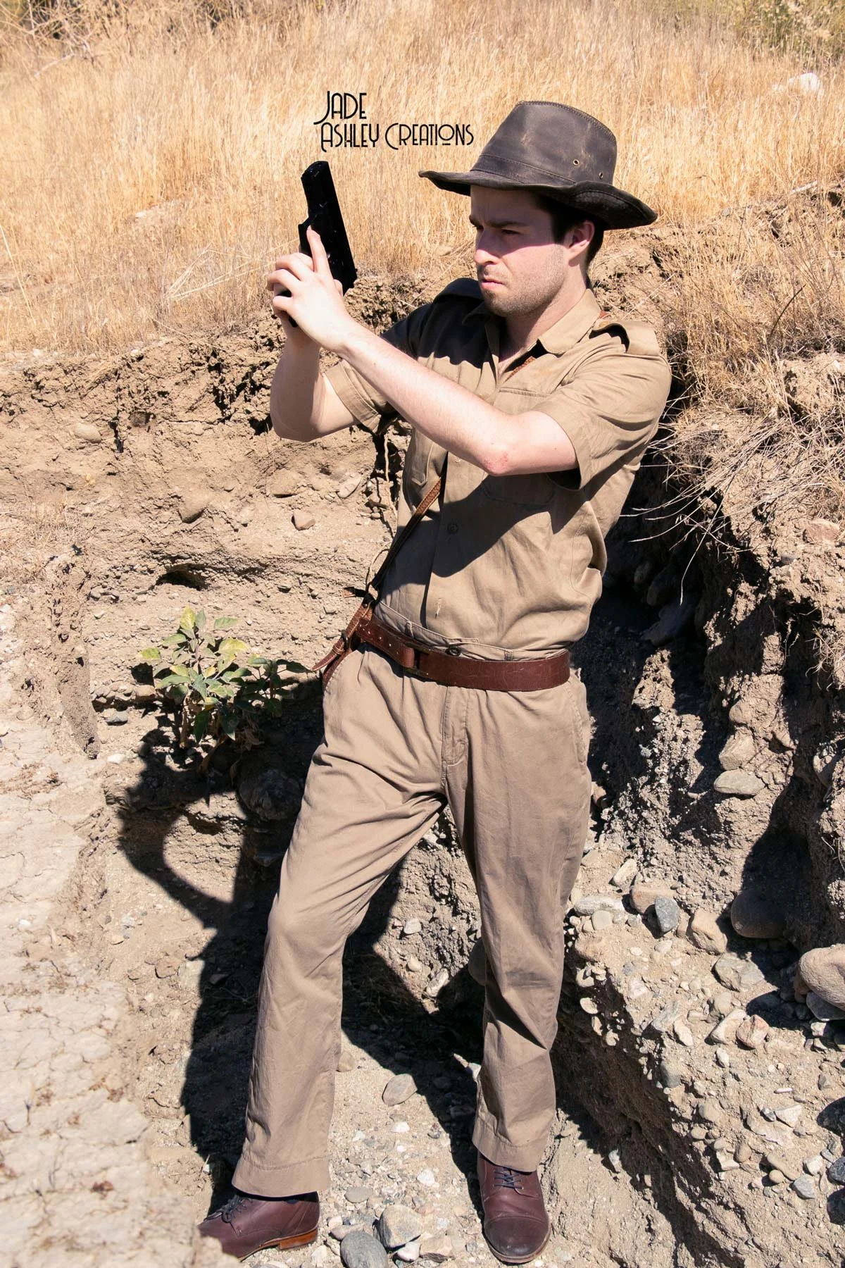 A man dressed as a park ranger stands in a dry, rocky outdoor area, aiming a handgun at an angle.