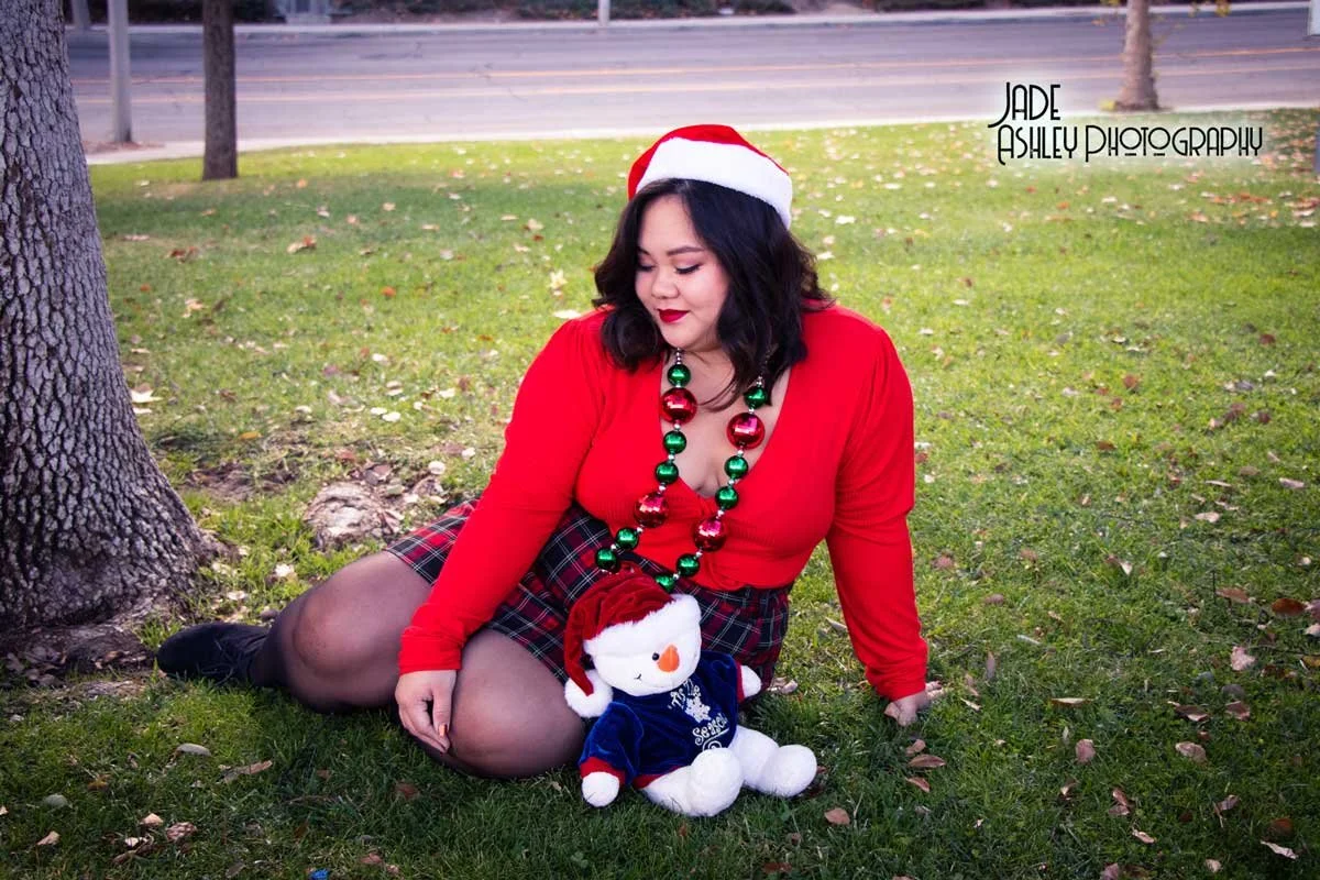 A woman in a red top, plaid skirt, black tights, and a Santa hat sitting on grass by a tree, holding a Christmas-themed teddy bear, with a Christmas necklace and holiday decorations, during daytime.