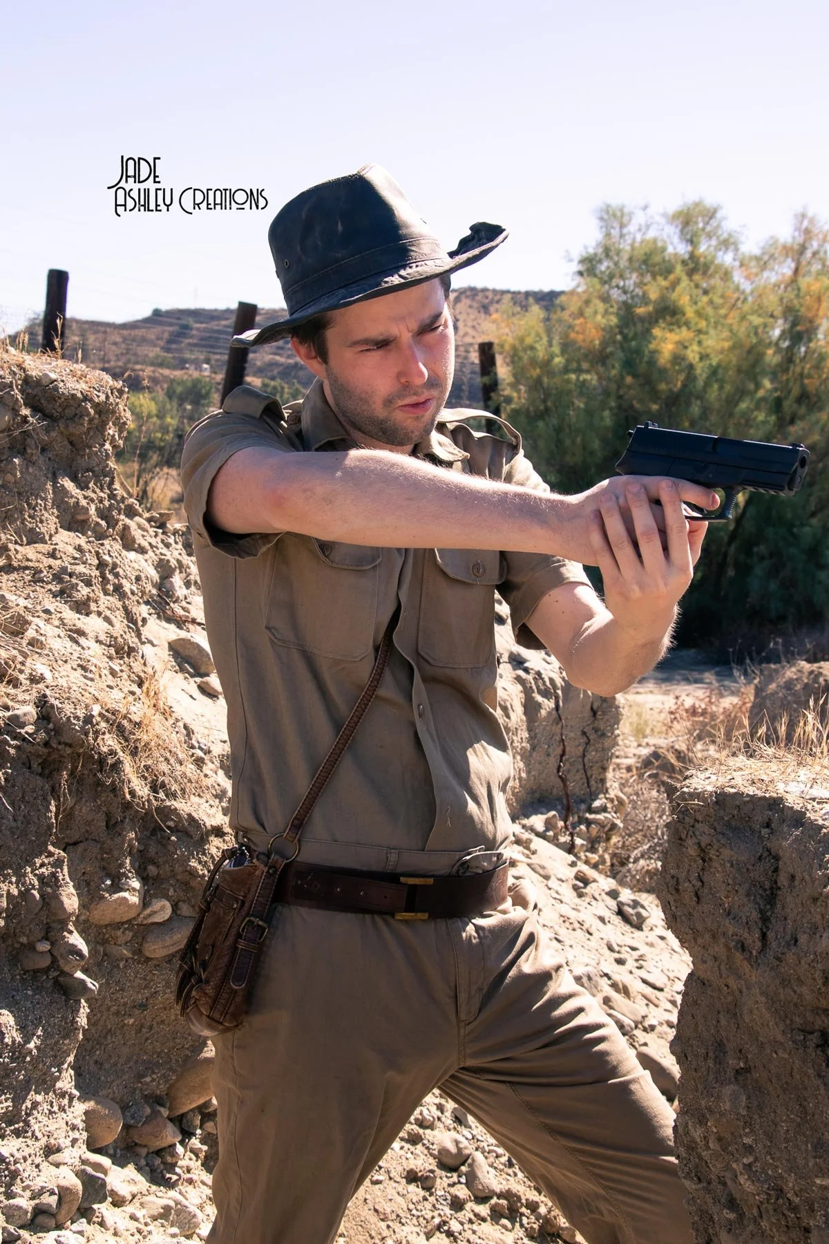 Man dressed as a park ranger aiming a handgun in a rocky outdoor landscape with trees and hills in the background.