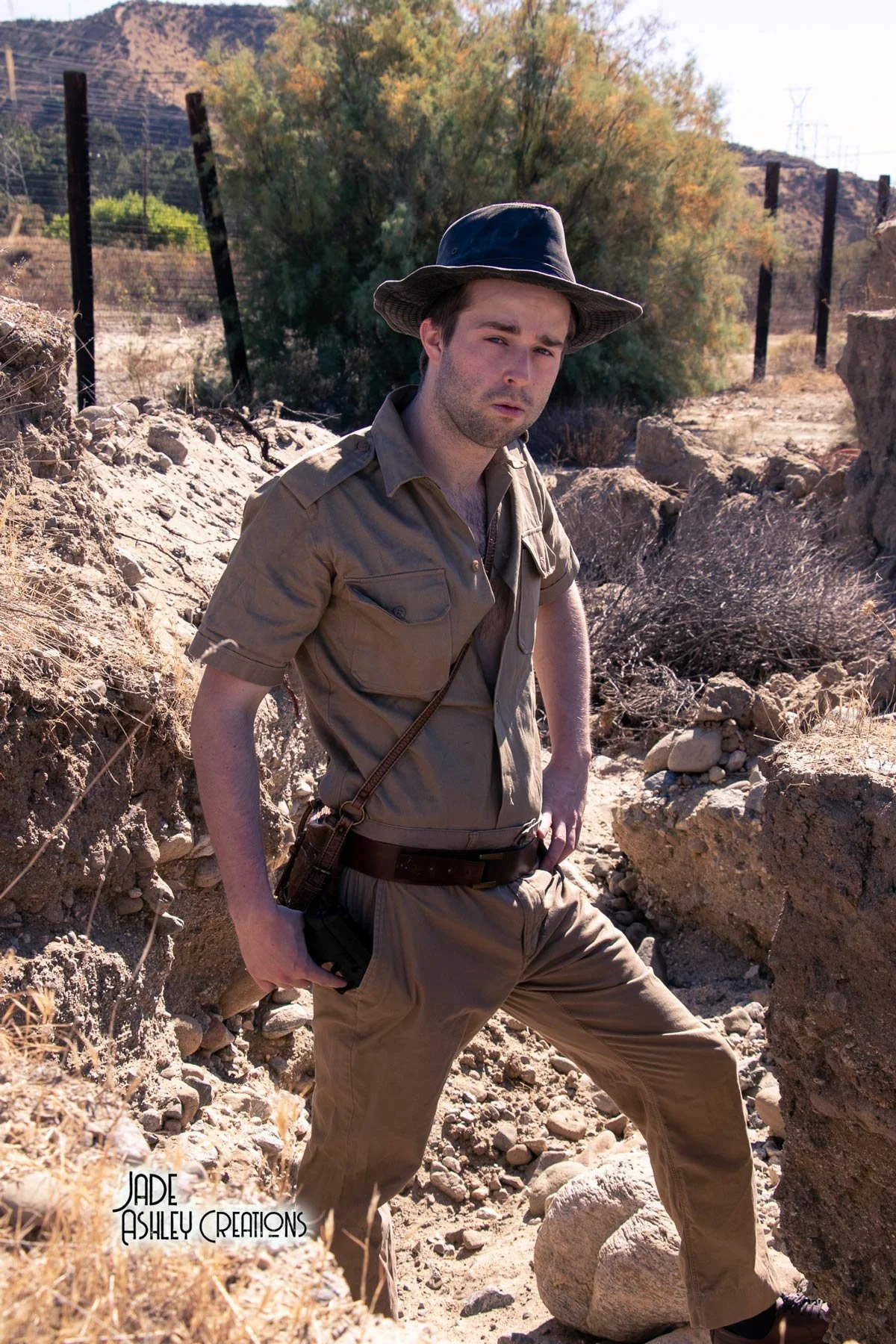 A man dressed as a park ranger or explorer standing in a rocky desert area with shrubs and trees in the background, wearing a wide-brimmed hat and khaki uniform.