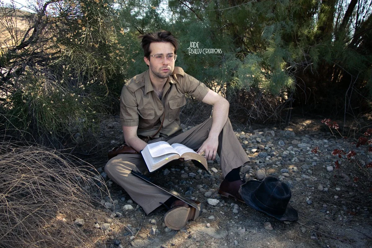 A young man with glasses and a brown shirt sitting on the ground among bushes, reading a book with a serious expression, with a black hat and a cane placed in front of him on rocky ground.
