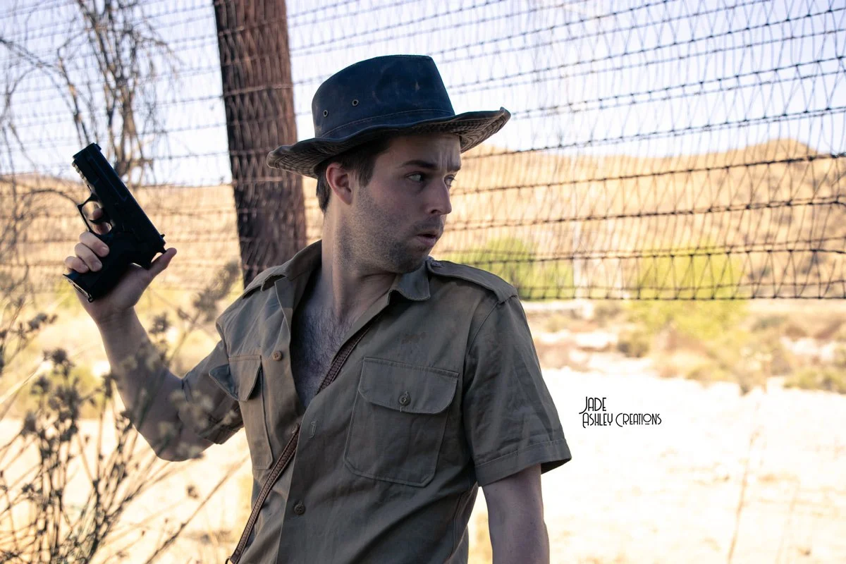 A man wearing a tan shirt and a wide-brimmed hat holding a handgun in an outdoor setting with barbed wire fencing and desert landscape in the background.