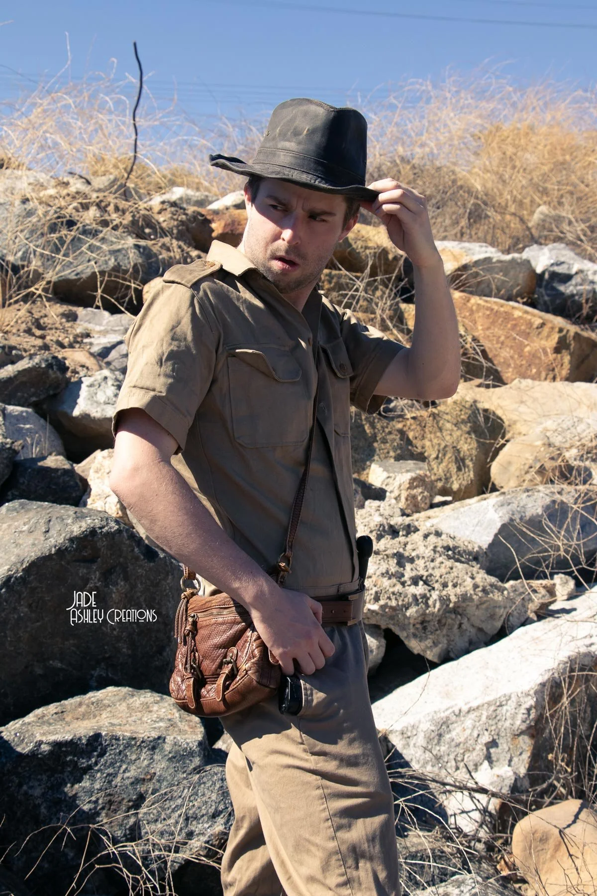 A man dressed in an explorer's outfit standing outdoors on rocky terrain with dry bushes, wearing a black hat, khaki shirt, and pants, holding a small brown bag and sunglasses, with a surprised expression.