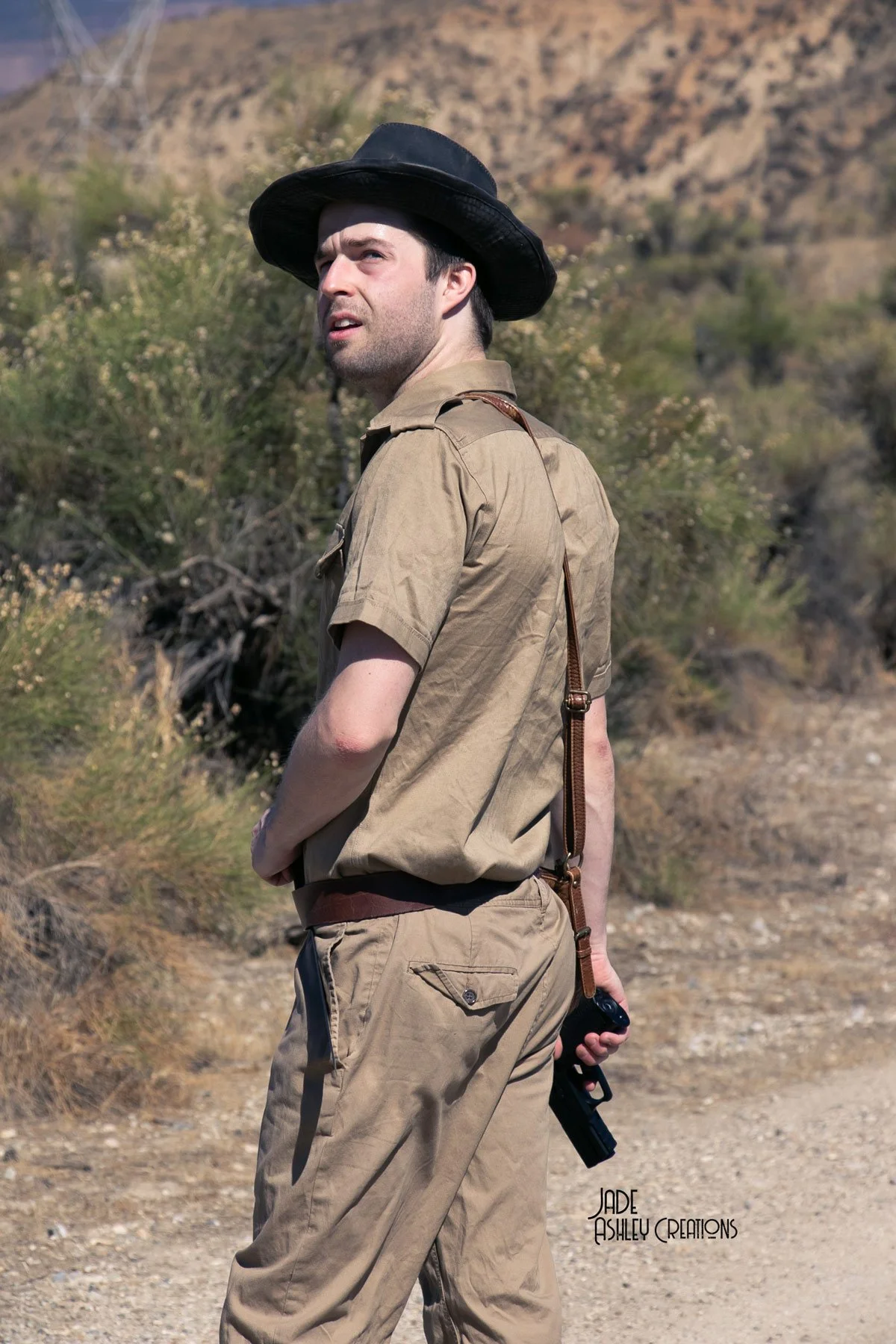 A man dressed as a park ranger holding a gun in a desert landscape with bushes and rocky hills.