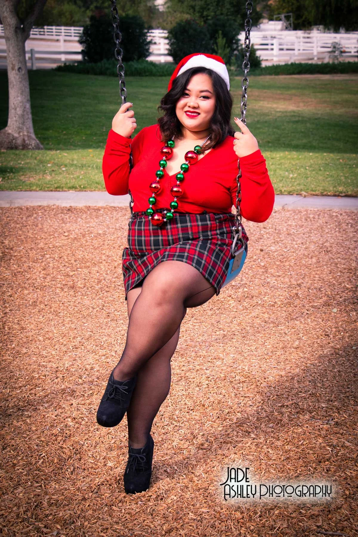 A woman wearing a Santa hat, red sweater, plaid skirt, black tights, and black shoes sitting on a playground swing, with Christmas beads around her neck and a park background.