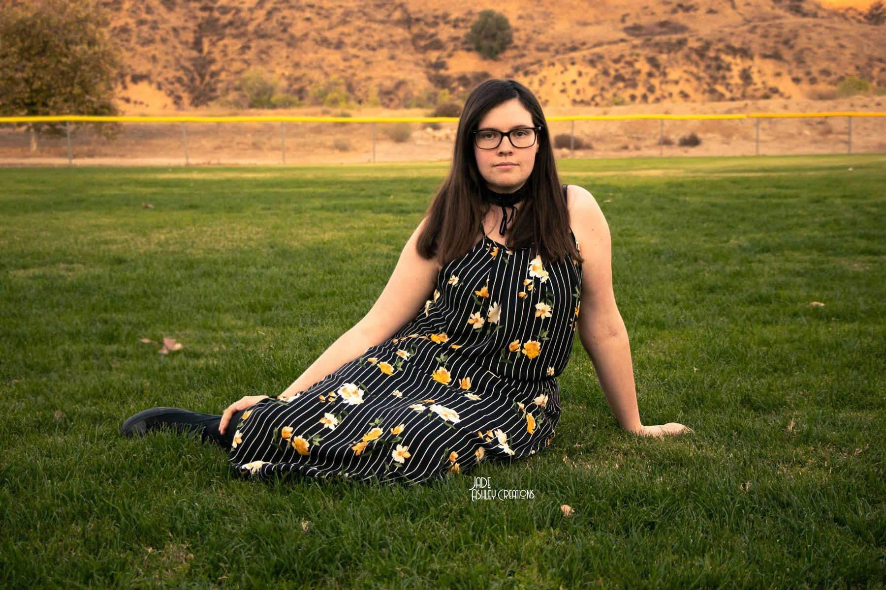 A woman with brown hair and glasses, wearing a black dress with white and yellow flowers, sitting on green grass in a park with hills and a baseball field in the background.