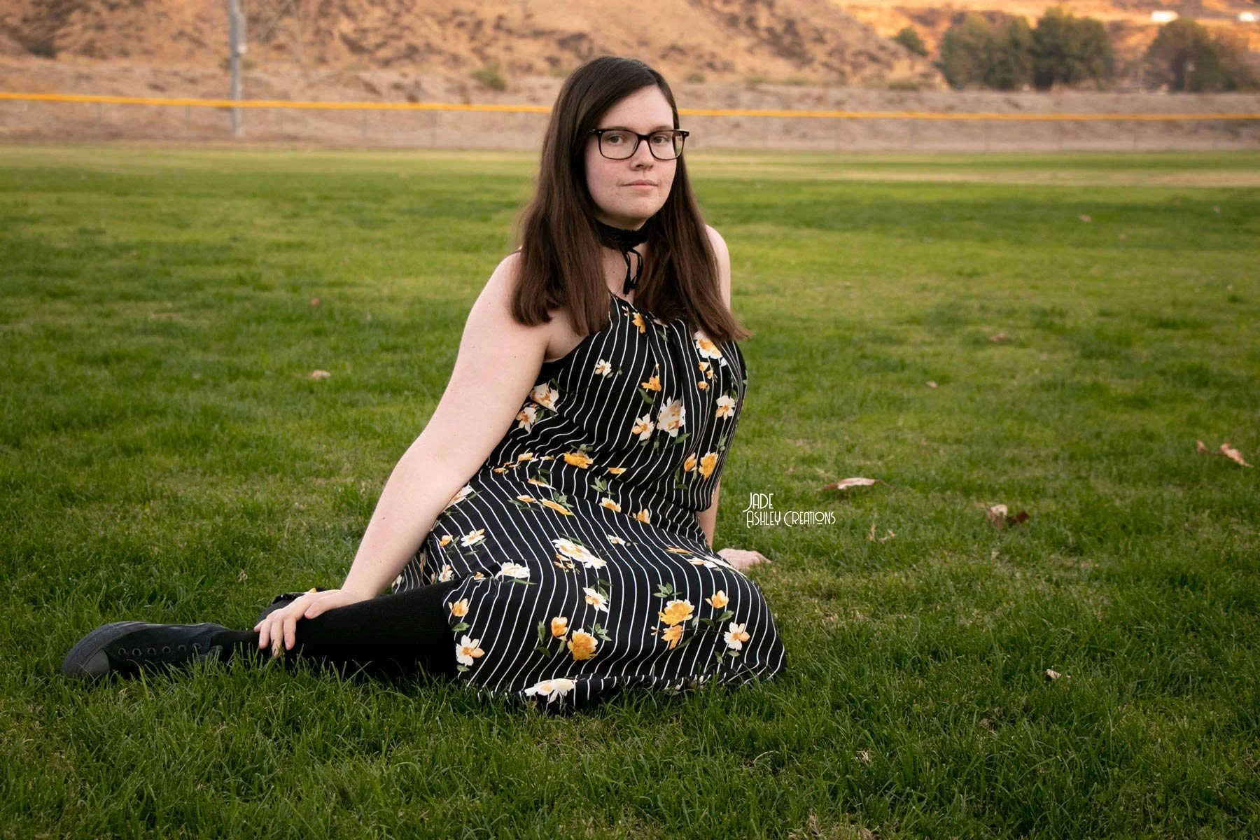 A young woman with long dark hair, glasses, and fair skin sitting on the grass field in a black sleeveless dress with white and yellow floral stripes. She is sitting with one hand on the ground and the other on her knee, looking at the camera with a 