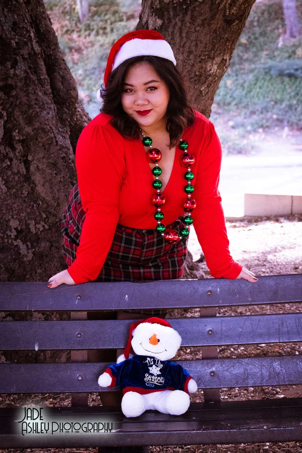 A woman dressed in a Christmas-themed red top with a plaid skirt, wearing a Santa hat and holiday bead necklace, sitting on a park bench with a plush snowman toy in front of her.