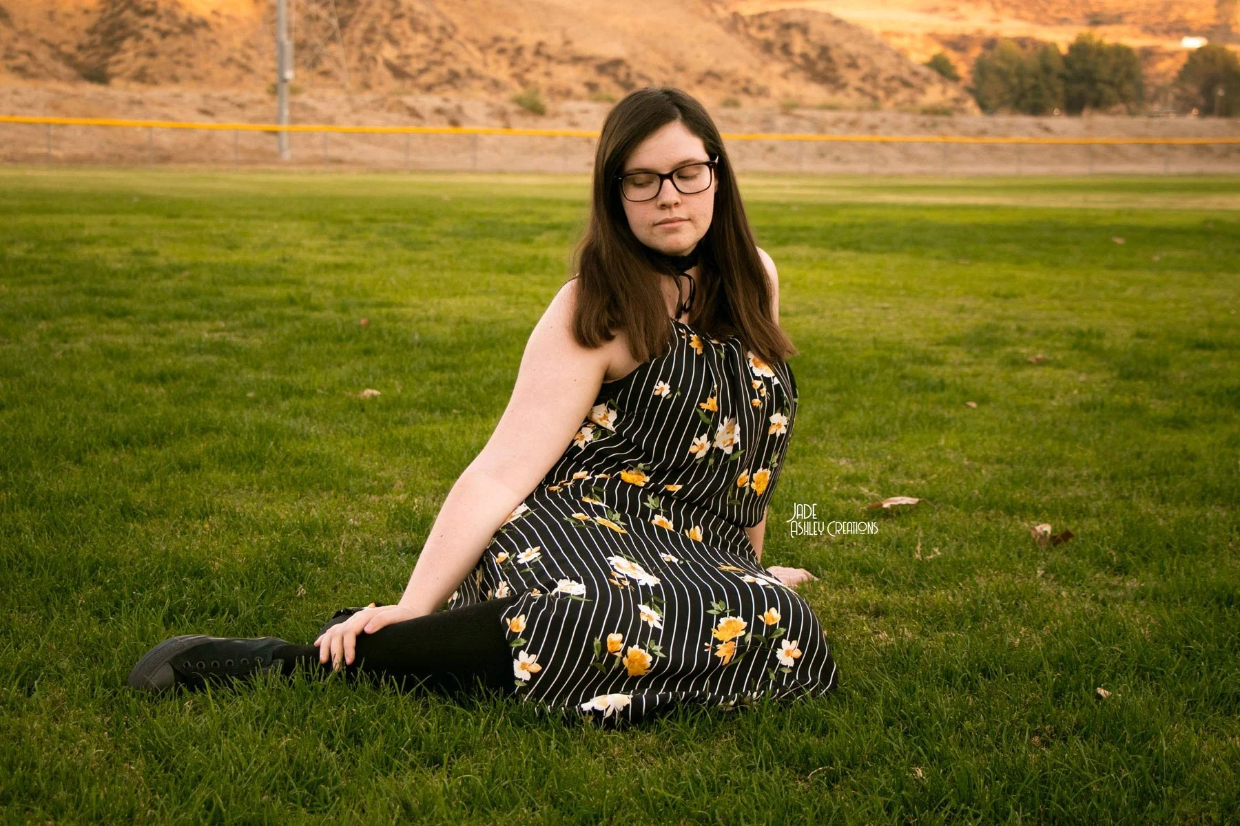 A young woman with glasses sitting on the grass in a kneeling pose with her eyes closed, wearing a black and white striped dress with a yellow floral pattern, in an open field with a fence and hills in the background.