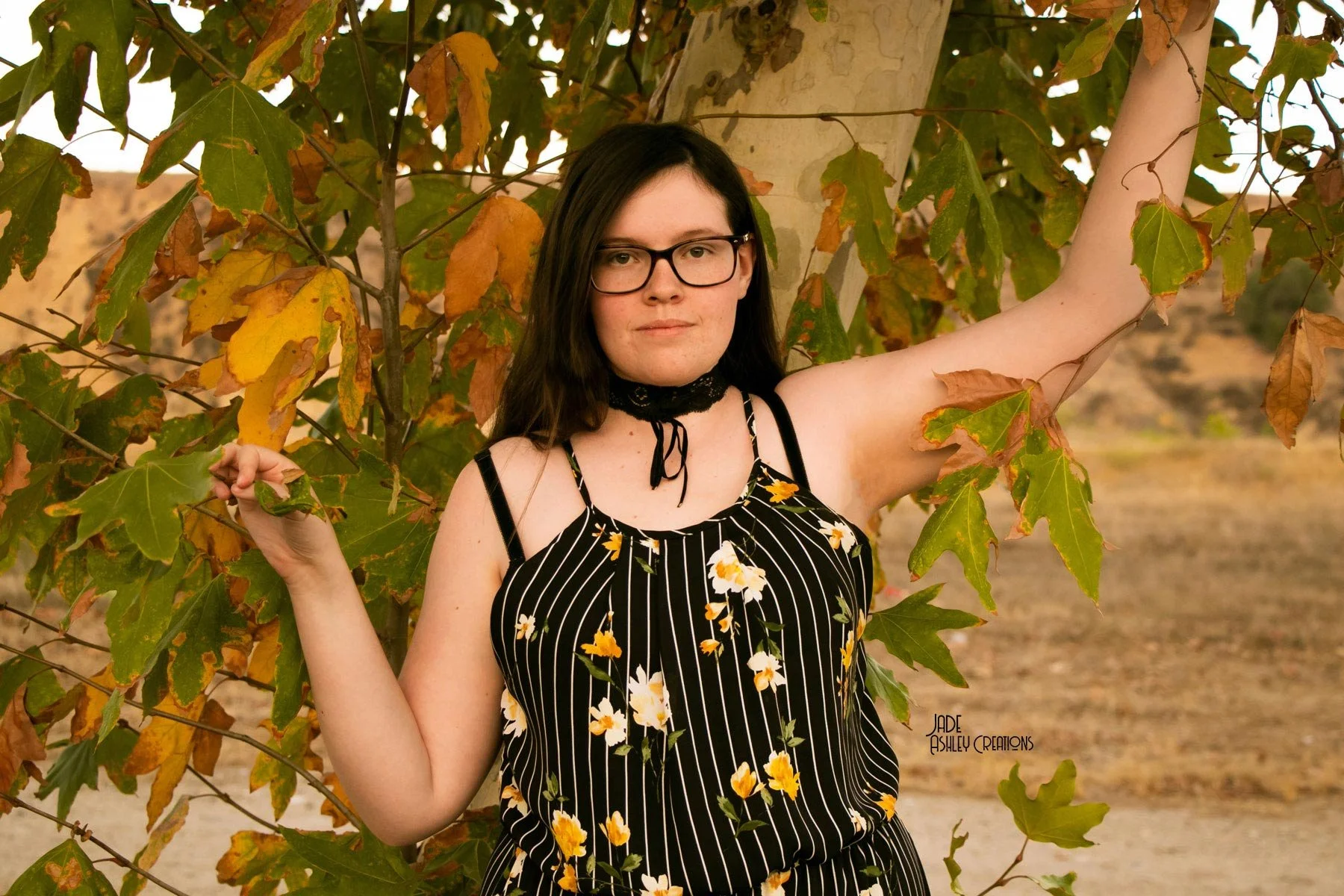 A young woman with dark hair and glasses is standing outdoors in front of a tree with green and yellow leaves. She is wearing a black and white striped dress with yellow and white flowers, and a black choker. Her right arm is raised and resting on a 