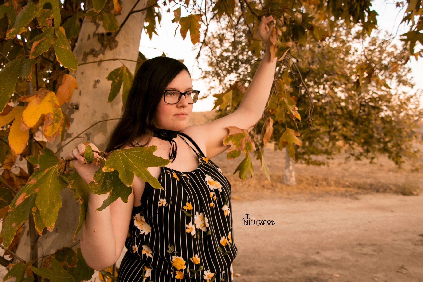A young woman with long dark hair, glasses, and wearing a black and white floral dress, stands outdoors near a tree with autumn leaves. She is holding a branch with large green leaves.