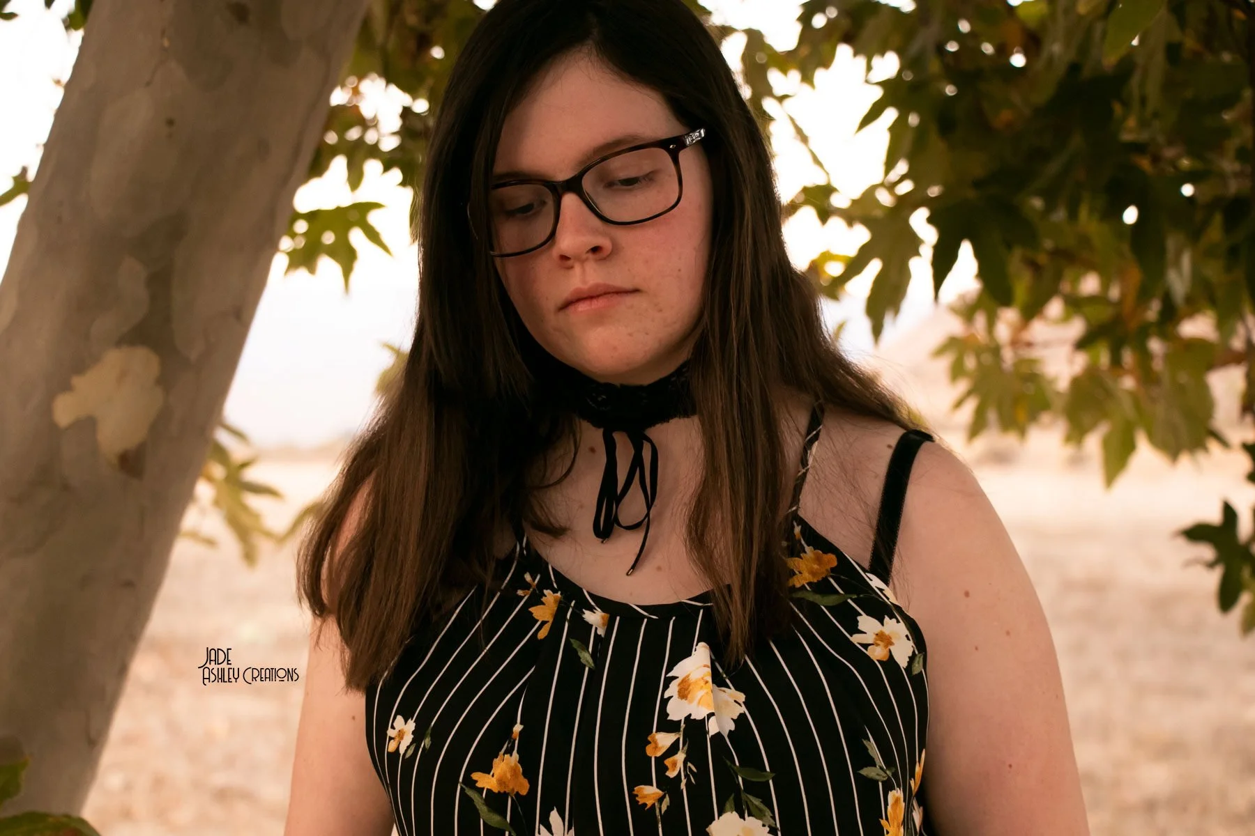 A young woman with long brown hair wearing black-rimmed glasses and a black dress with yellow and white floral pattern, standing outdoors near a tree with green leaves.